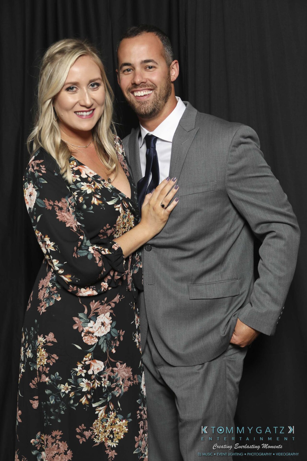 Megan-Matt-102718-Blow-WM-24 - TOMMY GATZ ENTERTAINMENT A woman in a floral dress and a man in a gray suit pose together, smiling in front of a dark backdrop.