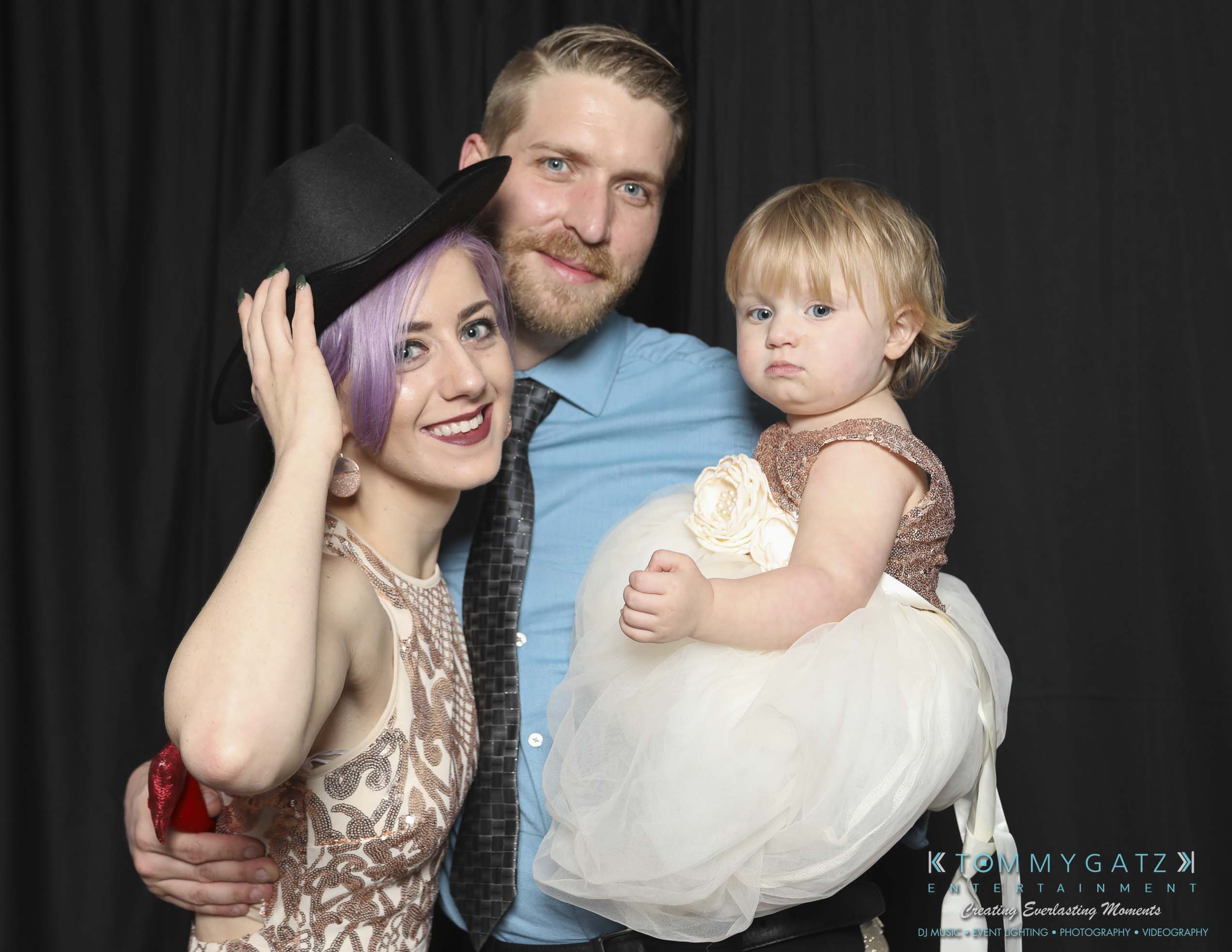 Megan-Matt-102718-Blow-WM-25 - TOMMY GATZ ENTERTAINMENT A woman with purple hair and a black hat, a man with a beard, and a toddler in a white dress pose together against a black background.