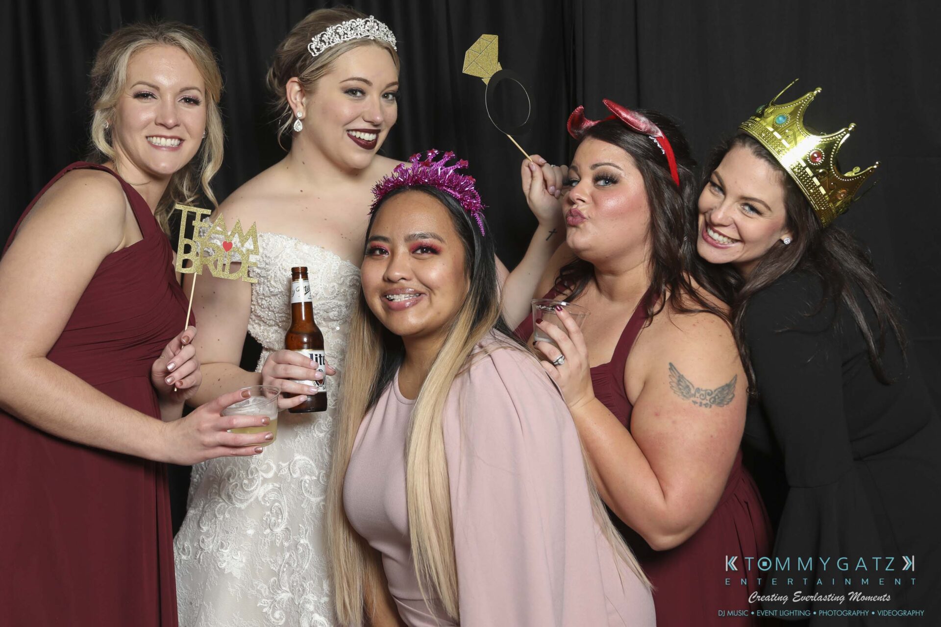 Megan-Matt-102718-Blow-WM-26 - TOMMY GATZ ENTERTAINMENT Five women posing together, one in a white dress holding a "Team Bride" sign and a beer bottle, others wearing crowns and playful accessories, all smiling.