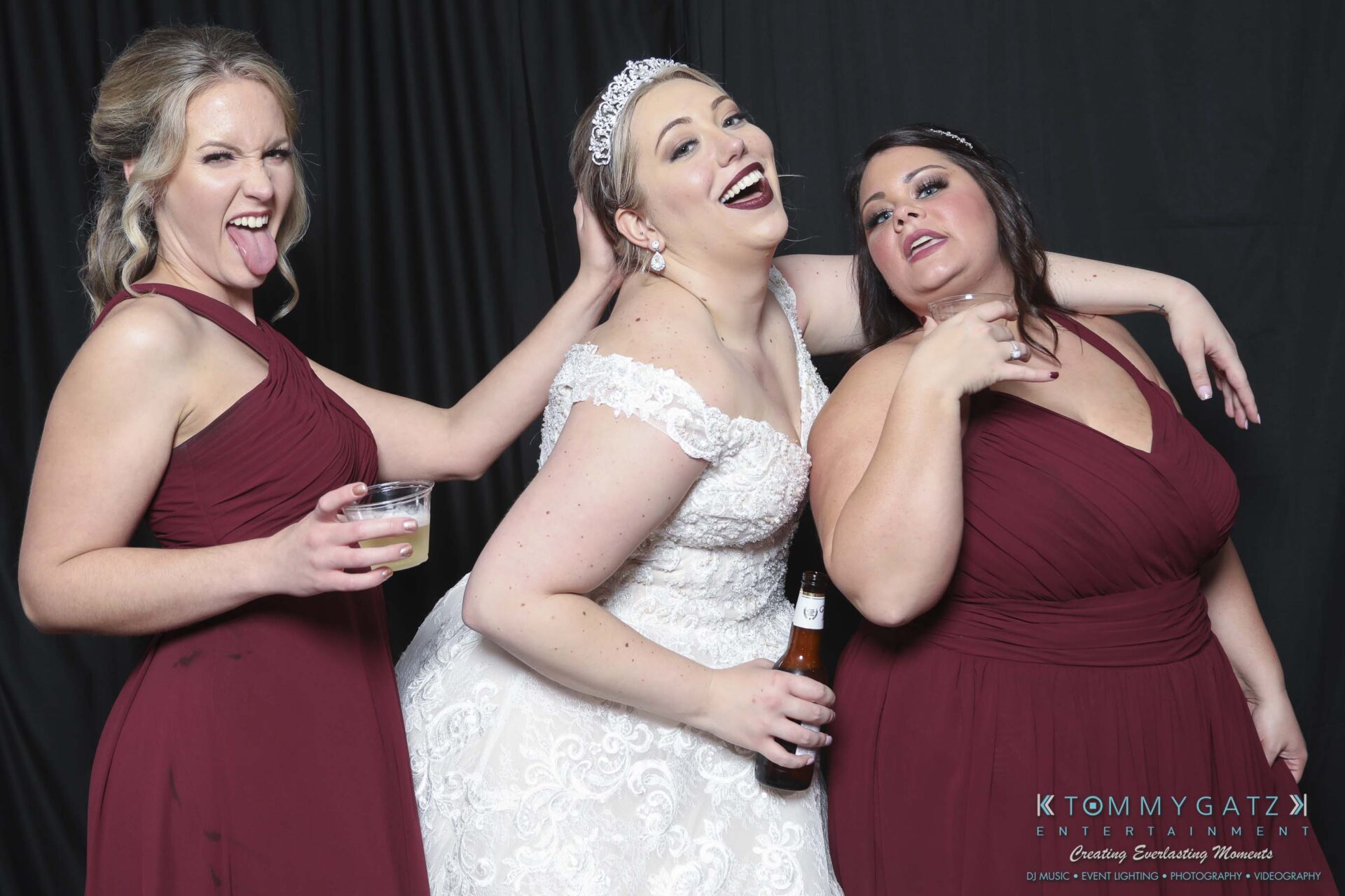 Megan-Matt-102718-Blow-WM-27 - TOMMY GATZ ENTERTAINMENT Three women in maroon dresses, one in a white bridal gown holding a beer, pose playfully against a dark backdrop.