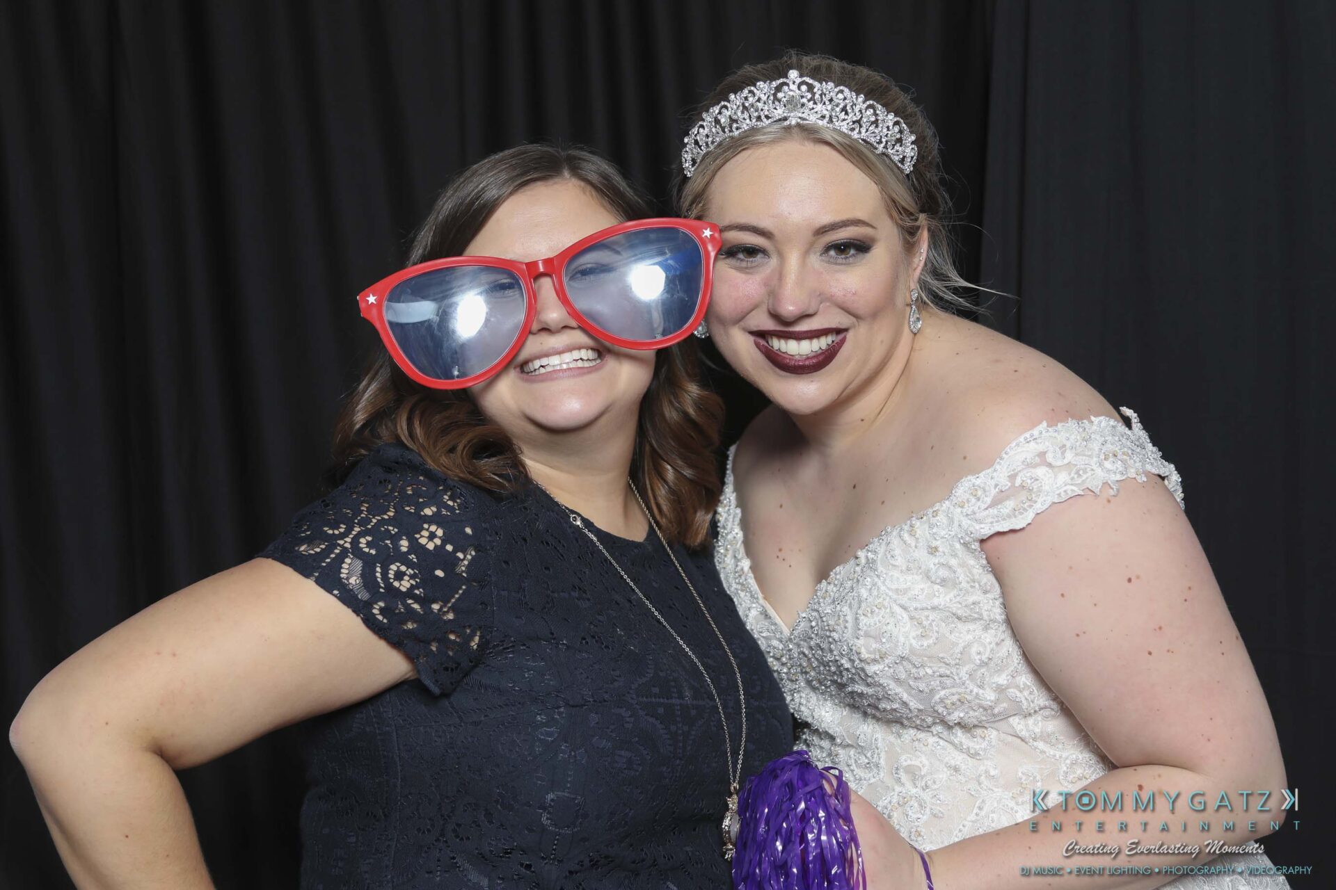 Megan-Matt-102718-Blow-WM-28 - TOMMY GATZ ENTERTAINMENT Two women posing in a photo booth. One wears oversized red glasses, and the other is in a white lace dress and tiara, holding a purple pom-pom.