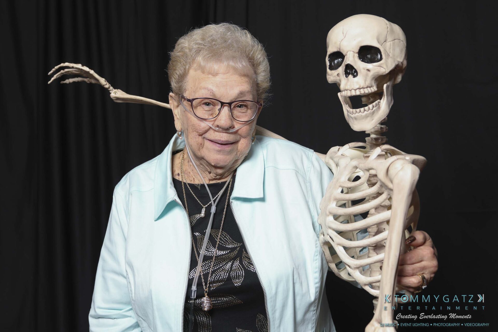 Megan-Matt-102718-Blow-WM-29 - TOMMY GATZ ENTERTAINMENT An elderly woman with glasses and a light blue jacket poses with a skeleton against a black backdrop.