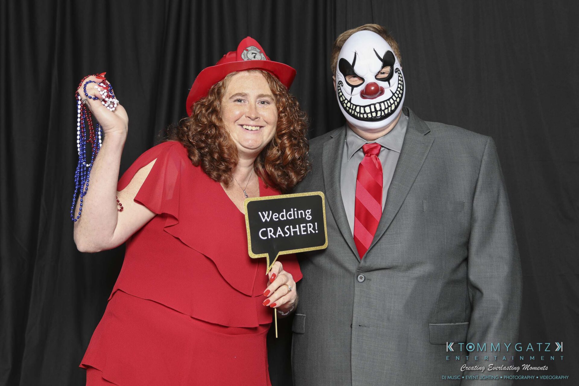 Megan-Matt-102718-Blow-WM-3 - TOMMY GATZ ENTERTAINMENT A woman in a red dress holds beads and a "Wedding Crasher!" sign, standing next to a person in a suit wearing a sinister clown mask.