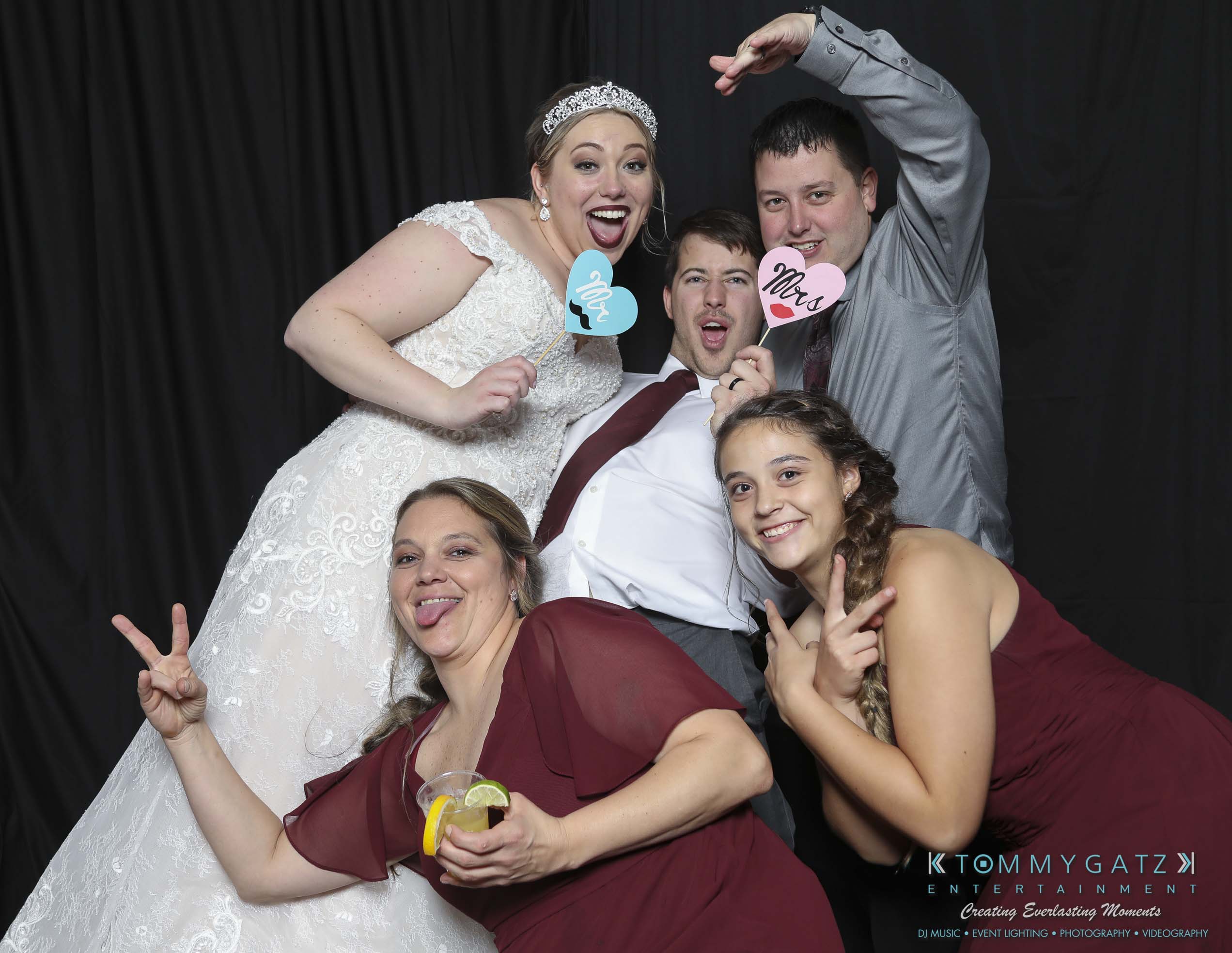 Megan-Matt-102718-Blow-WM-33 - TOMMY GATZ ENTERTAINMENT A group of five people posing at a photo booth. One person is in a wedding dress, and others hold cardboard props. Everyone is smiling or making funny faces against a dark backdrop.