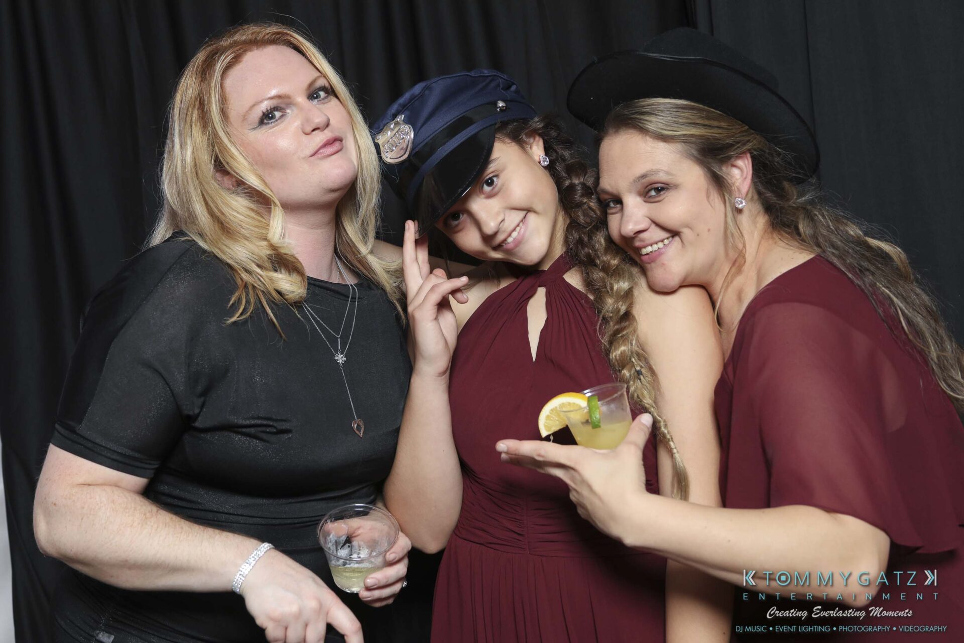 Megan-Matt-102718-Blow-WM-34 - TOMMY GATZ ENTERTAINMENT Three women pose together, one wearing a police hat, in front of a black backdrop. Two hold drinks, and all are smiling.