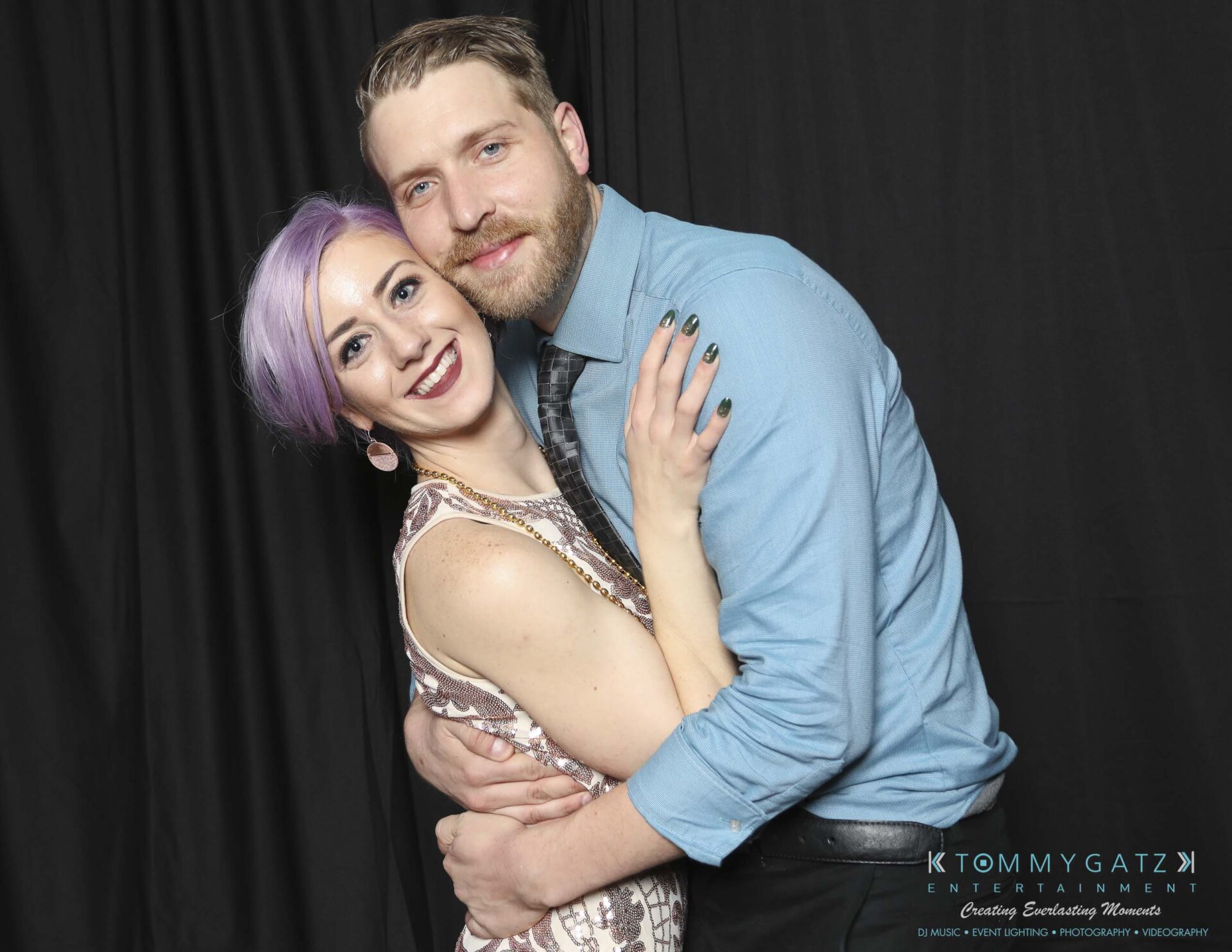 Megan-Matt-102718-Blow-WM-35 - TOMMY GATZ ENTERTAINMENT A couple poses together, smiling, in formal attire against a black backdrop. The woman has purple hair, and the man has light brown hair and a beard.