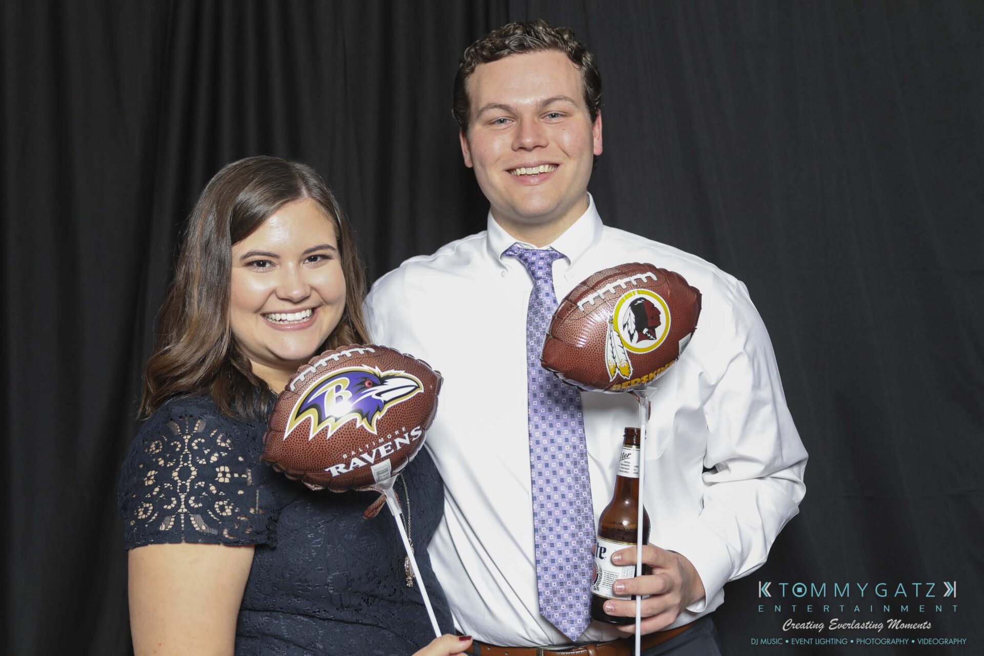 Megan-Matt-102718-Blow-WM-5 - TOMMY GATZ ENTERTAINMENT A man and woman smile while holding NFL-themed balloons, one with a Ravens logo and another with a Washington logo. The man holds a beer bottle. They stand in front of a black backdrop.