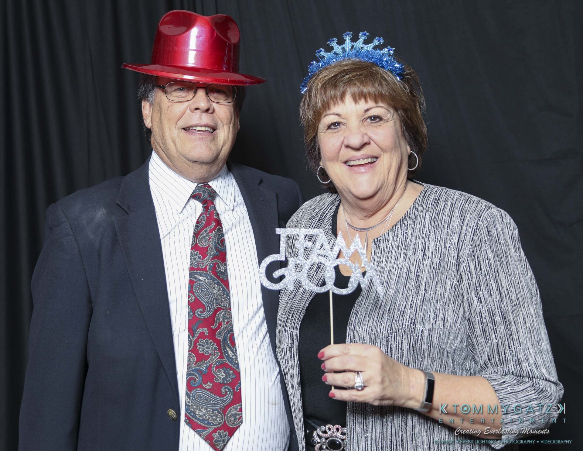 Megan-Matt-102718-Blow-WM-6 - TOMMY GATZ ENTERTAINMENT Two people smiling in a photo booth. The man wears a red hat and the woman holds a "Team Groom" sign, wearing a blue decorative headpiece.