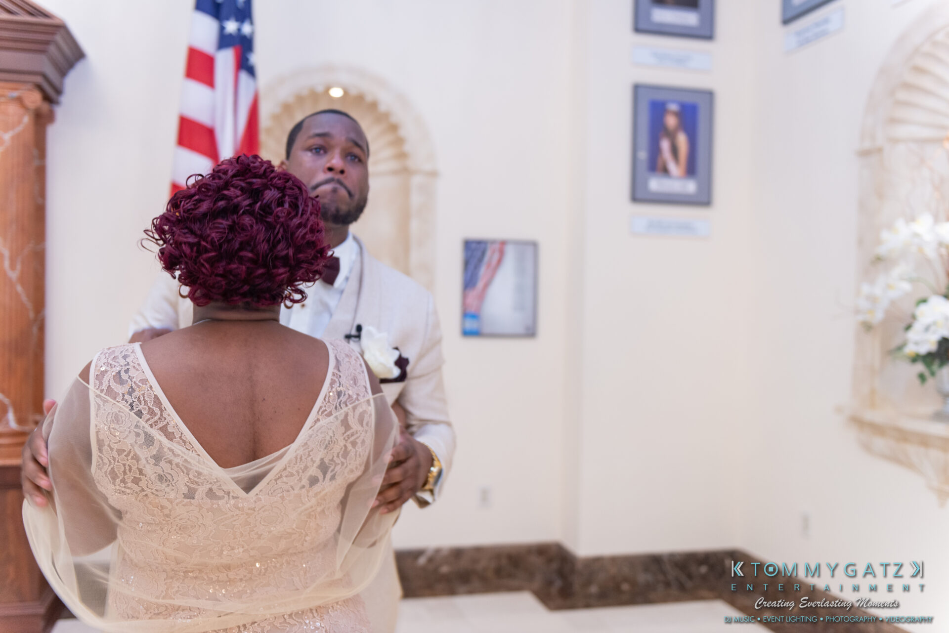 090119-Harris-Wedding-FINALS-Ceremony-Watermark-267 - TOMMY GATZ ENTERTAINMENT A couple dances together in a formal setting with an American flag and framed pictures on the wall.