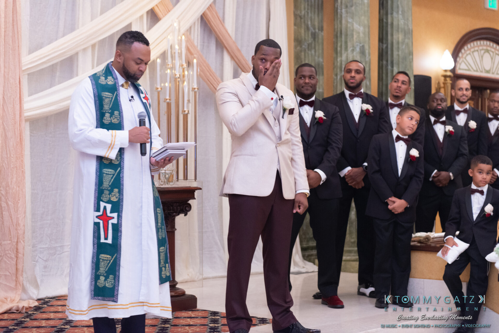 090119-Harris-Wedding-FINALS-Ceremony-Watermark-351 - TOMMY GATZ ENTERTAINMENT A groom wipes a tear during a wedding ceremony while a pastor reads. Groomsmen and boys in tuxedos stand nearby in a decorated room.