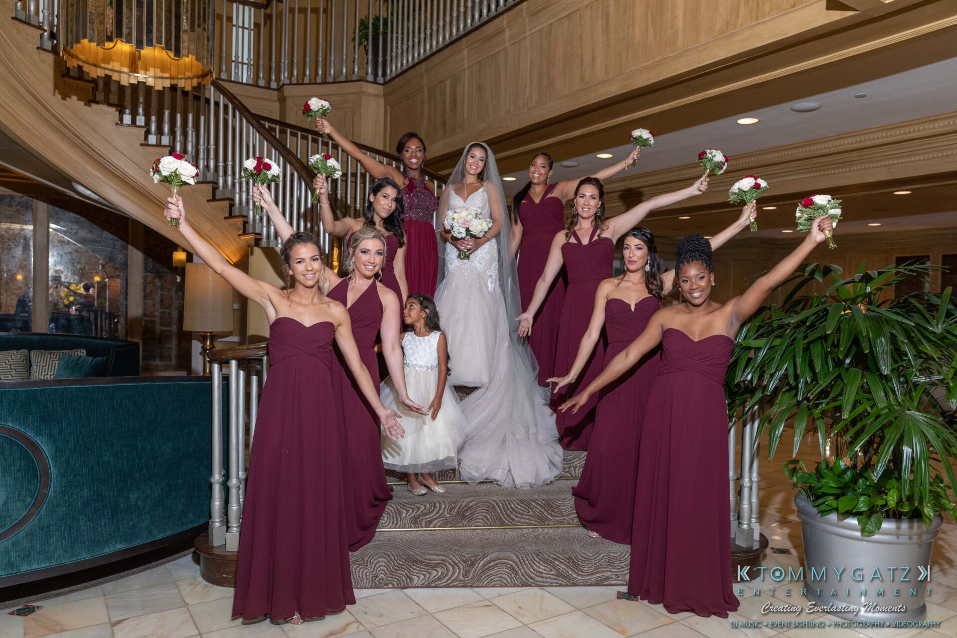 090119-Harris-Wedding-FINALS-Pre-Ceremony-Low-Res-WM-185 - TOMMY GATZ ENTERTAINMENT A bride and bridesmaids in burgundy dresses pose with bouquets on a staircase indoors.