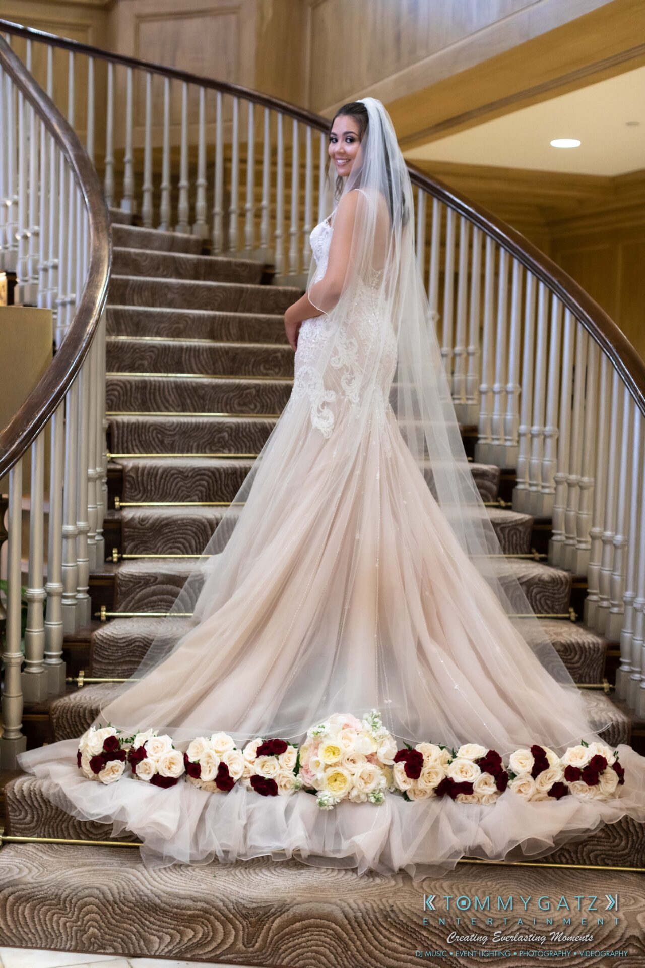090119-Harris-Wedding-FINALS-Pre-Ceremony-Low-Res-WM-222 - TOMMY GATZ ENTERTAINMENT Bride poses on a staircase in a lace wedding gown with a long train and veil, surrounded by floral arrangements.