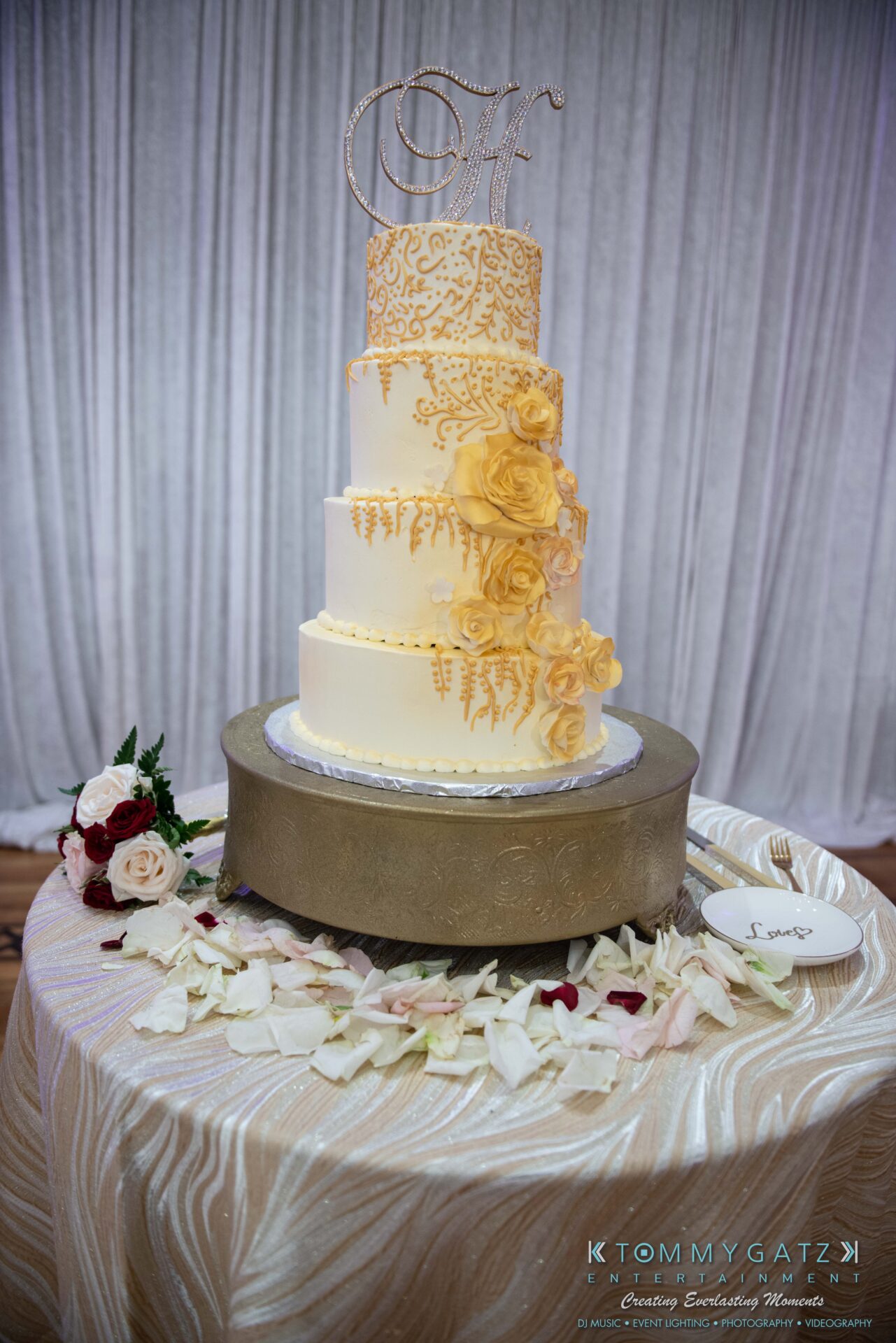 090119-Harris-Wedding-FINALS-Reception-Low-Res-WM-433 - TOMMY GATZ ENTERTAINMENT Four-tiered white and gold wedding cake with floral decorations on a round table adorned with rose petals and a small bouquet. The top features a monogram decoration.