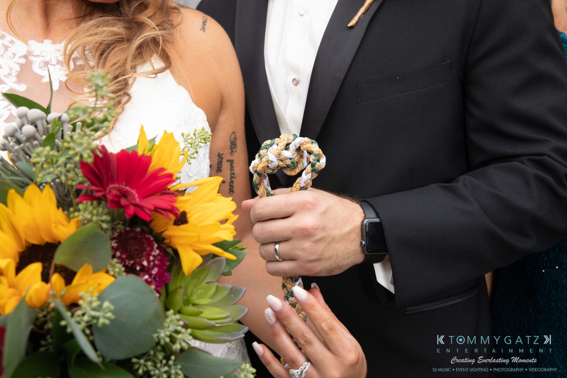 091419-Proctor-Wedding-FINALS-Formals-Low-Res-WM-468 - TOMMY GATZ ENTERTAINMENT A couple in wedding attire holds a braided cord. The bride has a bouquet with sunflowers and red flowers. The groom wears a black suit.