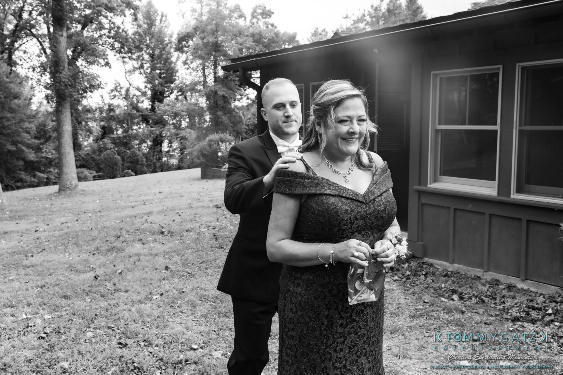 091419-Proctor-Wedding-FINALS-PreCeremony-Low-Res-WM-101 - TOMMY GATZ ENTERTAINMENT A man in a suit pins a corsage on a smiling woman in a dark dress outside a house, with trees in the background.