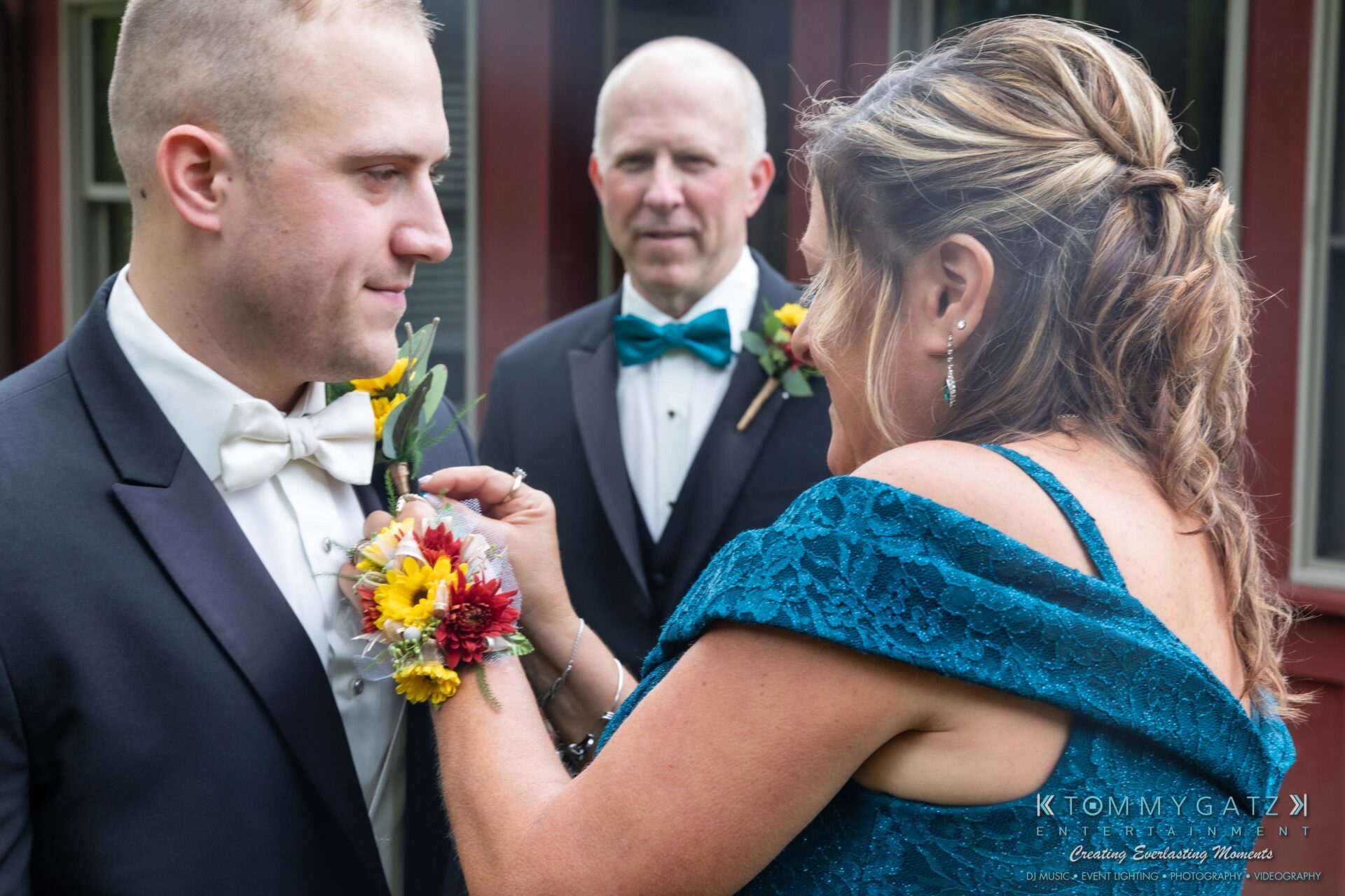 091419-Proctor-Wedding-FINALS-PreCeremony-Low-Res-WM-119 - TOMMY GATZ ENTERTAINMENT A woman adjusts the boutonniere of a man in a tuxedo, while another man stands in the background.