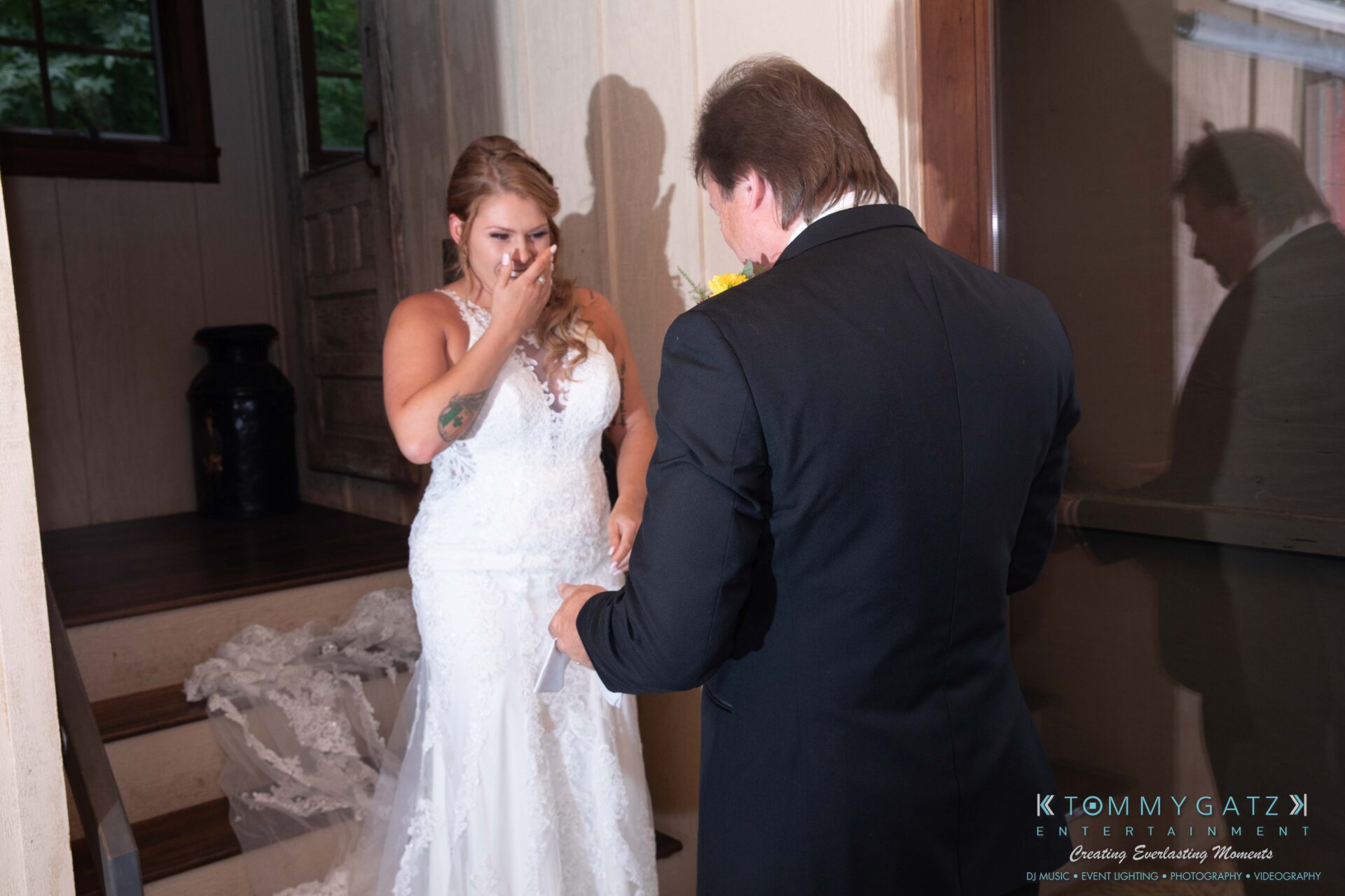 091419-Proctor-Wedding-FINALS-PreCeremony-Low-Res-WM-149 - TOMMY GATZ ENTERTAINMENT Bride in a white dress stands near stairs, covering her mouth with her hand, facing a man in formal attire. They appear emotional in an indoor setting.