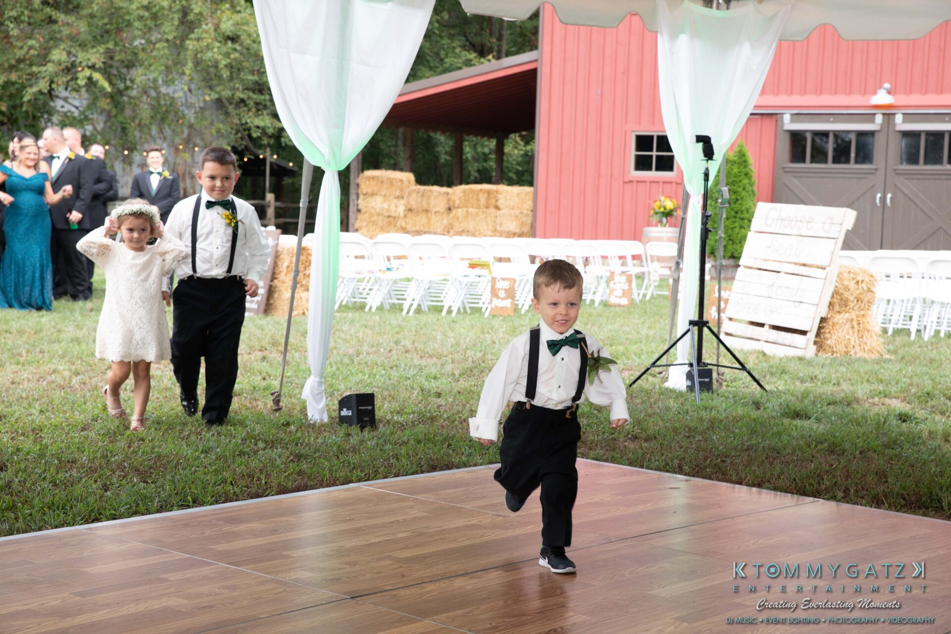 091419-Proctor-Wedding-FINALS-Reception-Low-Res-WM-545 - TOMMY GATZ ENTERTAINMENT Three children in formal attire run joyfully on a wooden dance floor outdoors, with a group of people and a red barn in the background.