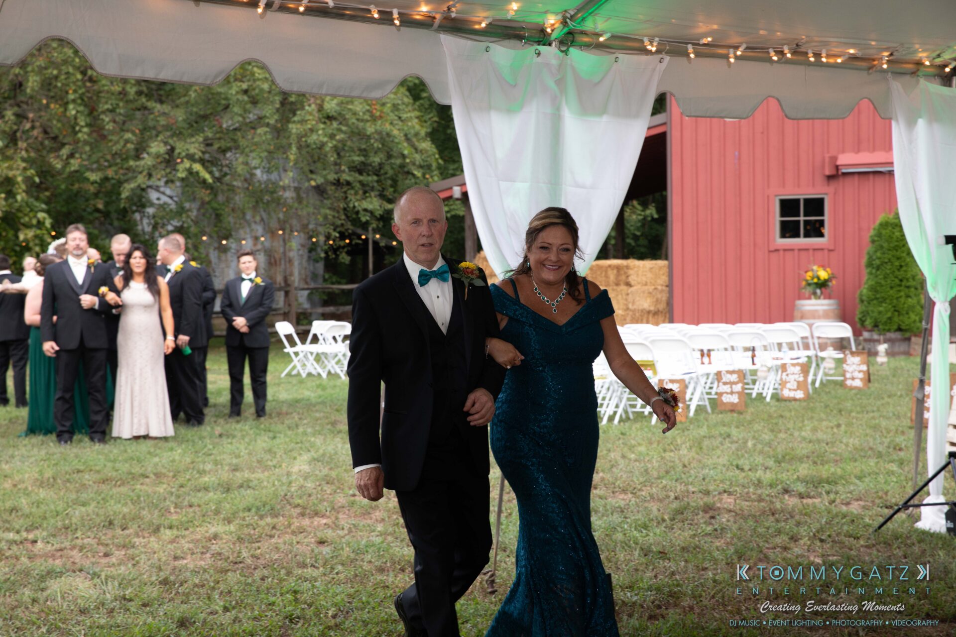 091419-Proctor-Wedding-FINALS-Reception-Low-Res-WM-549 - TOMMY GATZ ENTERTAINMENT A man and woman in formal attire walk under a tent at an outdoor event. A group of people and a red barn are visible in the background.