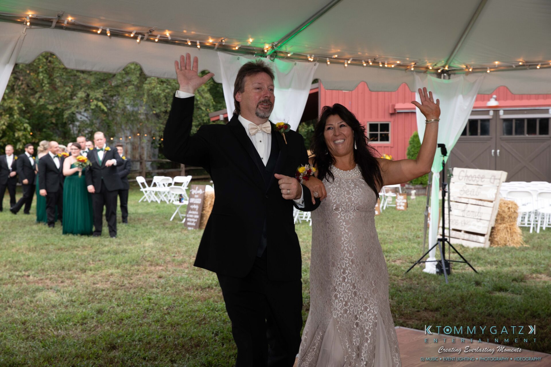 091419-Proctor-Wedding-FINALS-Reception-Low-Res-WM-554 - TOMMY GATZ ENTERTAINMENT A couple in formal attire enters an outdoor event tent, smiling and waving, with guests and greenery in the background.