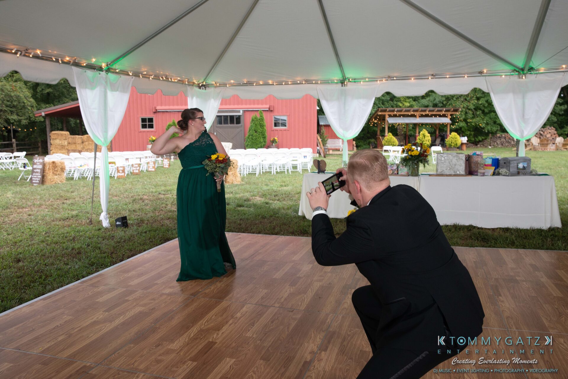 091419-Proctor-Wedding-FINALS-Reception-Low-Res-WM-558 - TOMMY GATZ ENTERTAINMENT A woman in a green dress poses with a bouquet while a man in a suit photographs her under a tent at a rustic outdoor event.