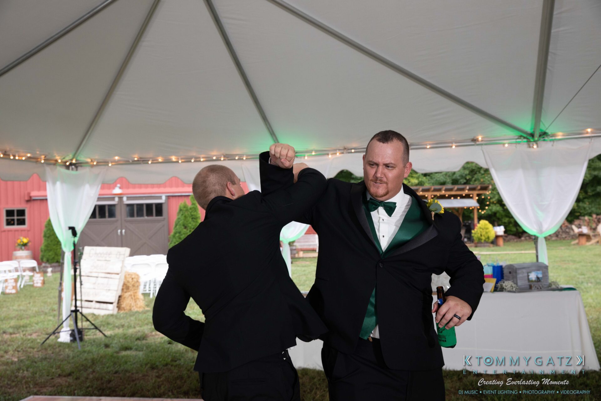 091419-Proctor-Wedding-FINALS-Reception-Low-Res-WM-562 - TOMMY GATZ ENTERTAINMENT Two men in suits high-fiving under a white tent at an outdoor event, with a barn and decorations in the background.