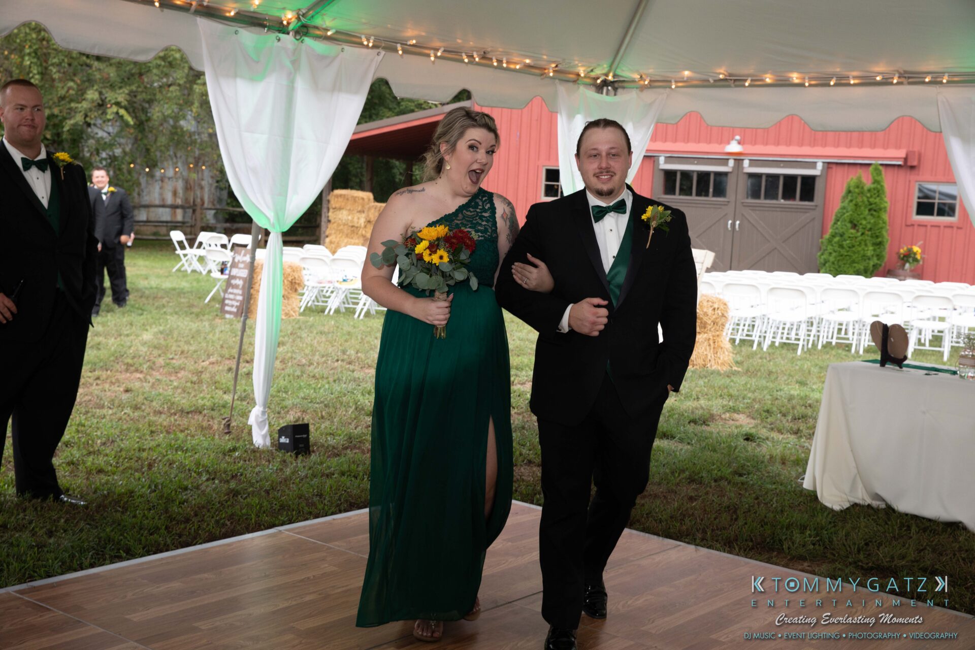 091419-Proctor-Wedding-FINALS-Reception-Low-Res-WM-566 - TOMMY GATZ ENTERTAINMENT A woman in a green dress and a man in a black suit with a green vest walk arm-in-arm under a tent at an outdoor event. Tables and chairs are arranged in the background.