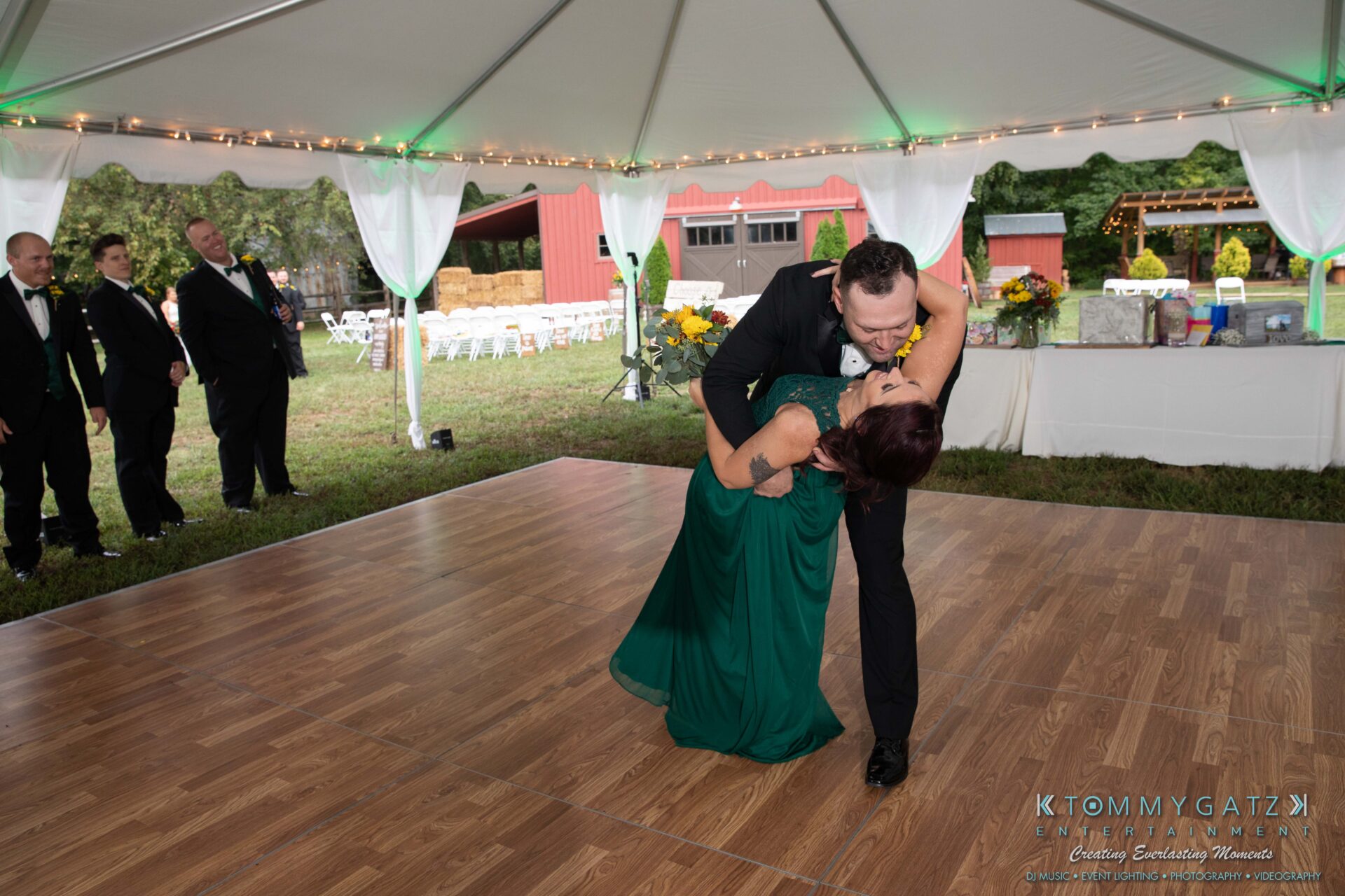 091419-Proctor-Wedding-FINALS-Reception-Low-Res-WM-569 - TOMMY GATZ ENTERTAINMENT A couple dances under a tent on a wooden floor at an outdoor event. Tables with flowers and a red barn are in the background.