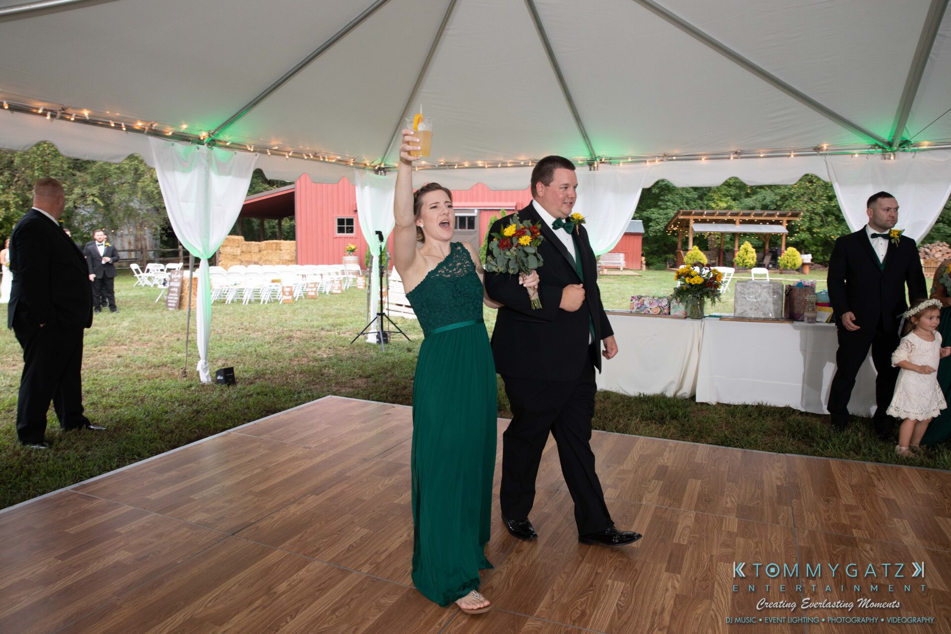 091419-Proctor-Wedding-FINALS-Reception-Low-Res-WM-573 - TOMMY GATZ ENTERTAINMENT A woman in a green dress raises a glass while holding a bouquet next to a man in a suit under a tent at an outdoor event.