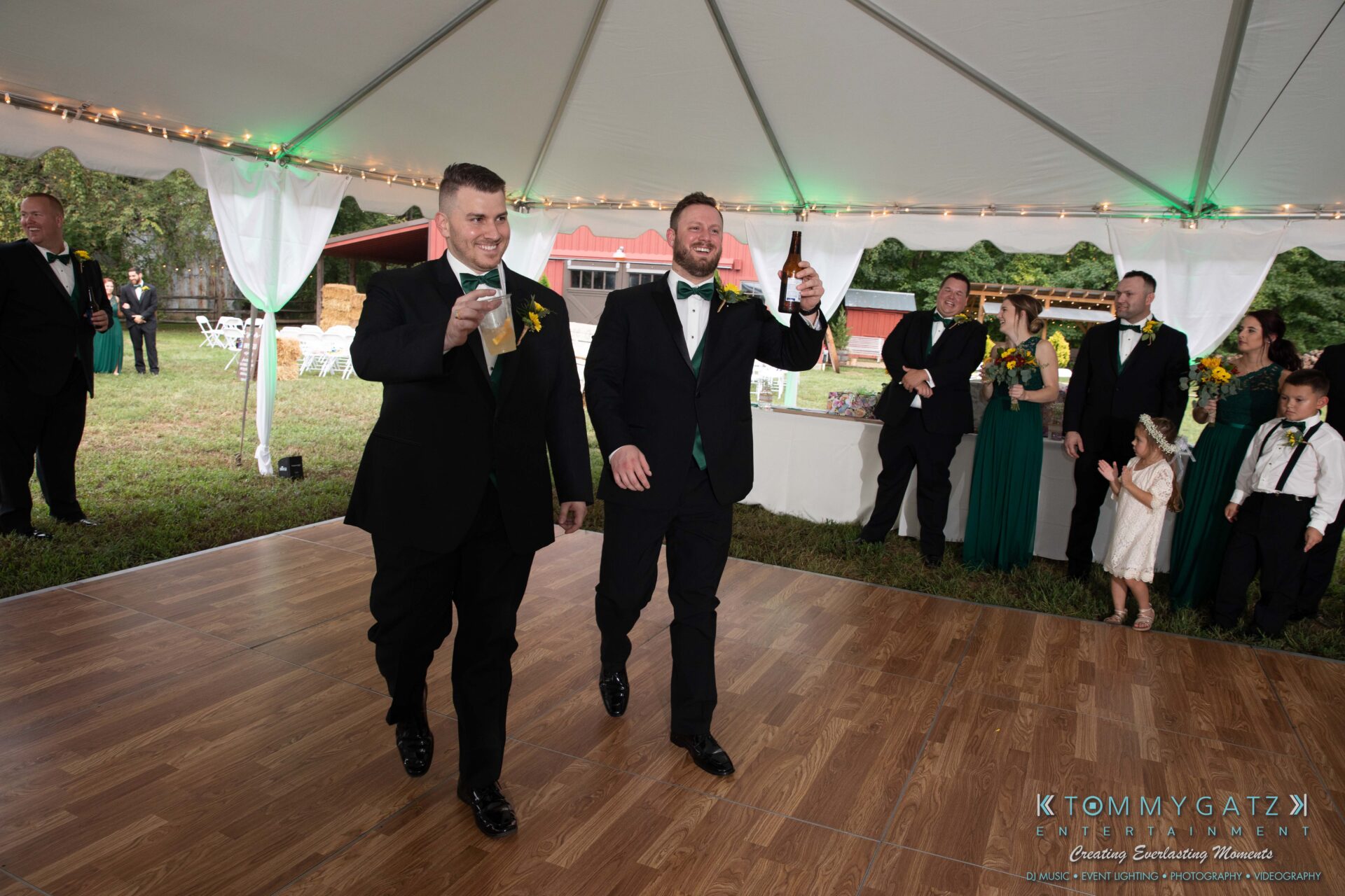 091419-Proctor-Wedding-FINALS-Reception-Low-Res-WM-574 - TOMMY GATZ ENTERTAINMENT Two men in suits enter a tented outdoor event holding drinks. Guests, some in formal attire and others casual, stand around the perimeter. A dance floor and grassy area are visible.