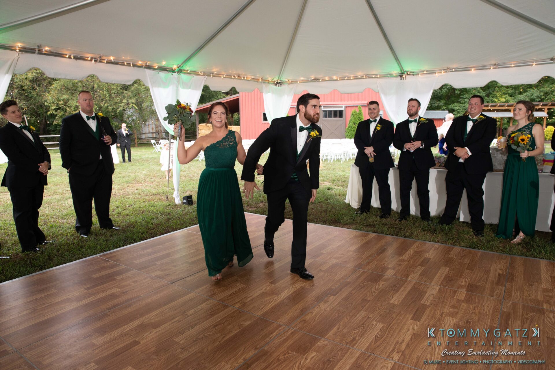 091419-Proctor-Wedding-FINALS-Reception-Low-Res-WM-575 - TOMMY GATZ ENTERTAINMENT A couple in formal attire enters a tented dance floor, holding hands. Five people stand in the background, dressed for an event.