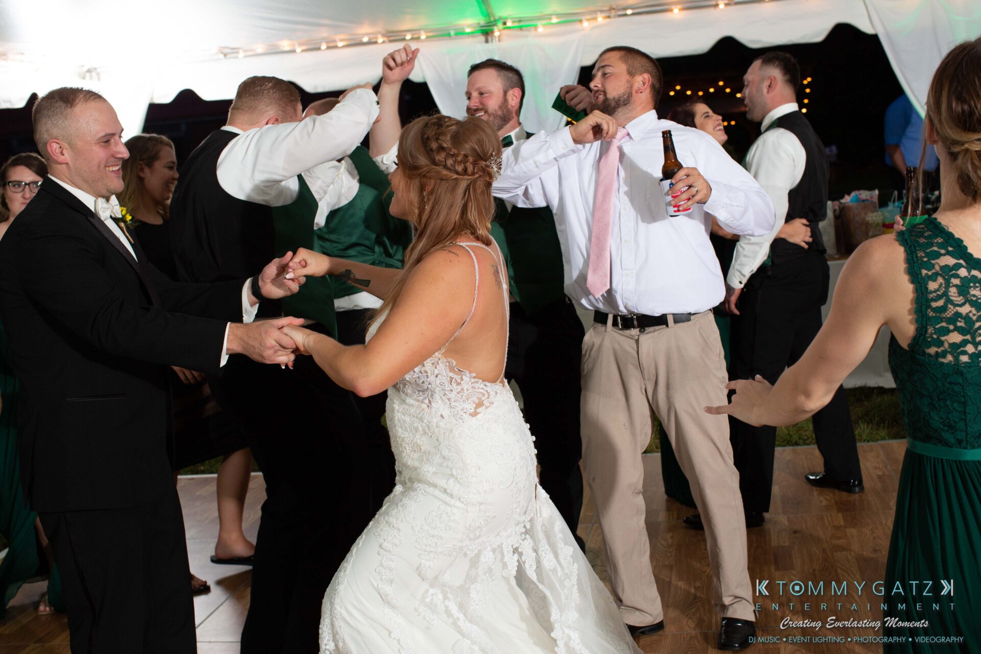 091419-Proctor-Wedding-FINALS-Reception-Low-Res-WM-717 - TOMMY GATZ ENTERTAINMENT A wedding reception with guests dancing energetically under a tent. A bride in a white gown dances with a guest in formal attire.