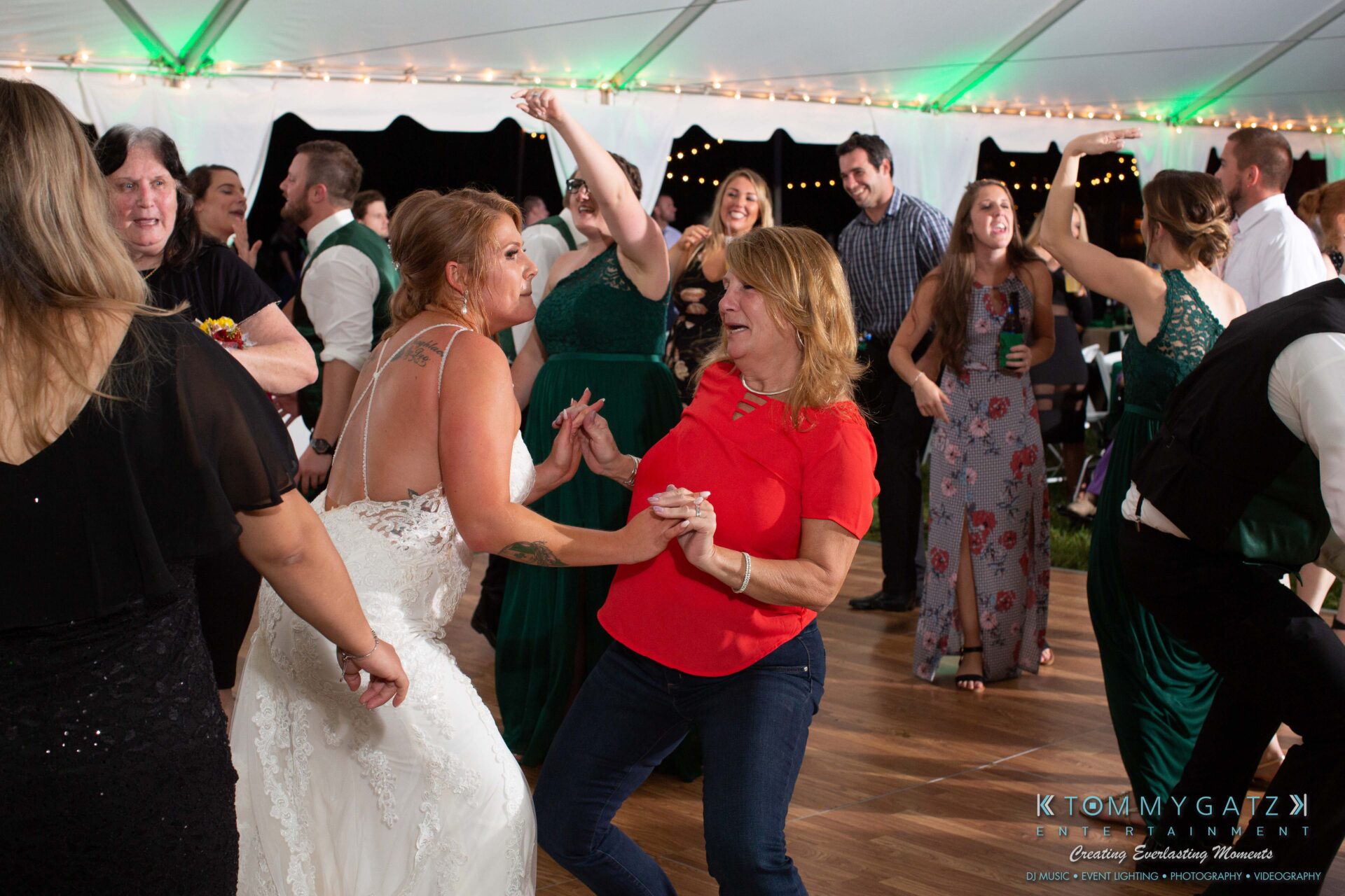 091419-Proctor-Wedding-FINALS-Reception-Low-Res-WM-735 - TOMMY GATZ ENTERTAINMENT People dancing energetically at a wedding reception under a tent with string lights. A woman in a wedding dress and another in a red top are in the foreground.