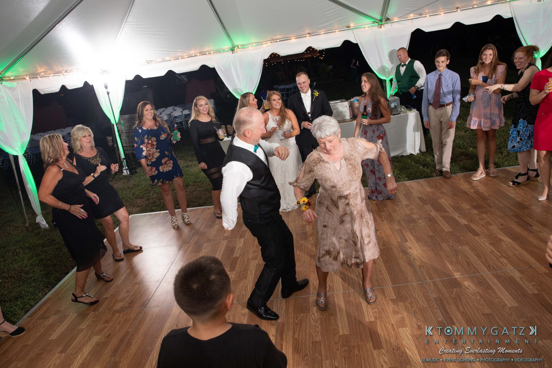 091419-Proctor-Wedding-FINALS-Reception-Low-Res-WM-758 - TOMMY GATZ ENTERTAINMENT People dancing on a wooden floor under a white tent at an outdoor event. A woman in a floral dress and a man in a vest are leading the dance in the foreground.