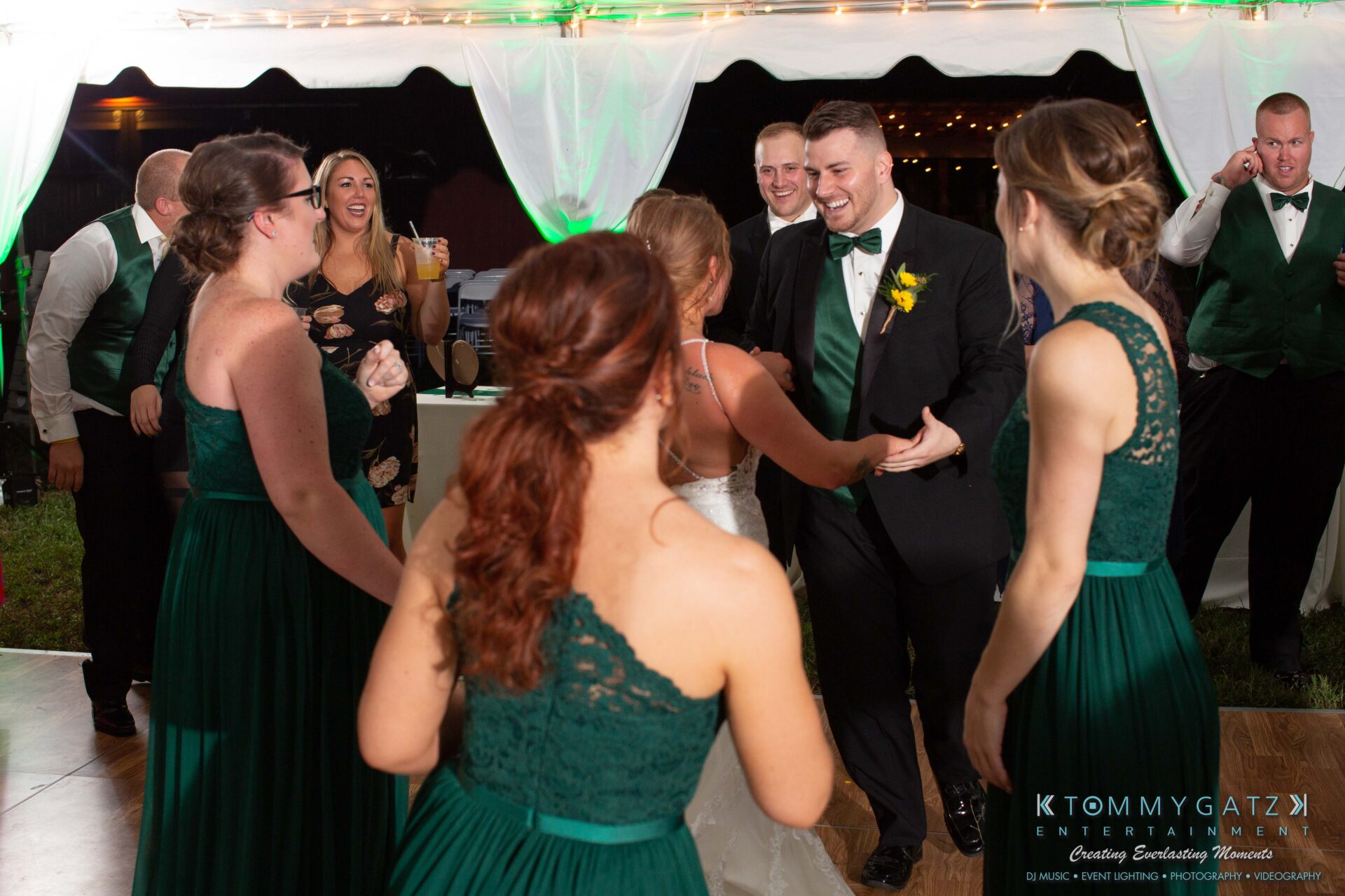 091419-Proctor-Wedding-FINALS-Reception-Low-Res-WM-765 - TOMMY GATZ ENTERTAINMENT A bride and groom dance joyfully with bridesmaids and groomsmen in green dresses and vests, under a tent with white drapery.