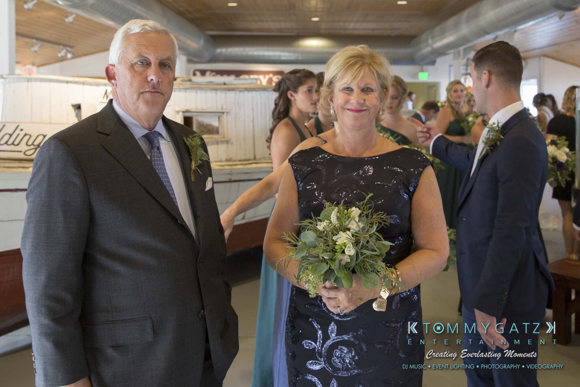Amanda-Tyler-102117-CEREMONY-Low-Res-WM-1 - TOMMY GATZ ENTERTAINMENT A man in a suit and a woman in a black dress holding flowers stand indoors with people in formal attire in the background.