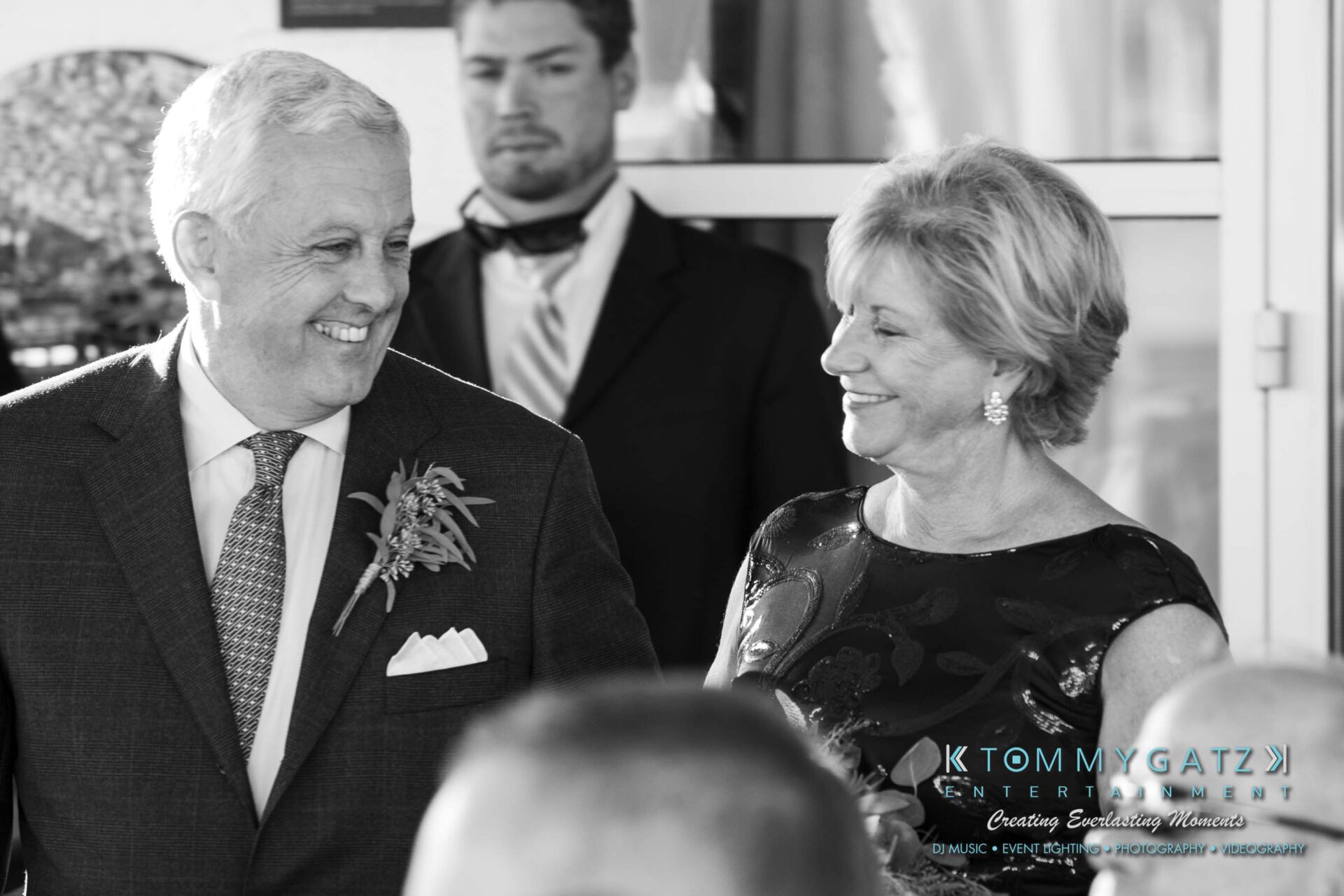 An older couple smiling at each other, dressed formally with a man in the background.