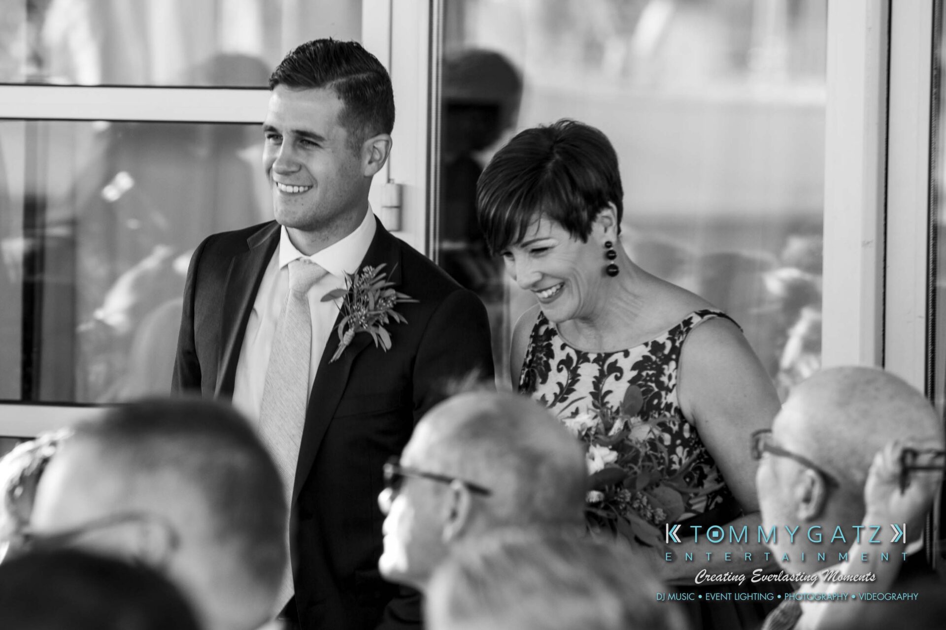 A man in a suit and a woman in a dress are standing and smiling at a gathering. They are near a glass door, with others seated nearby.