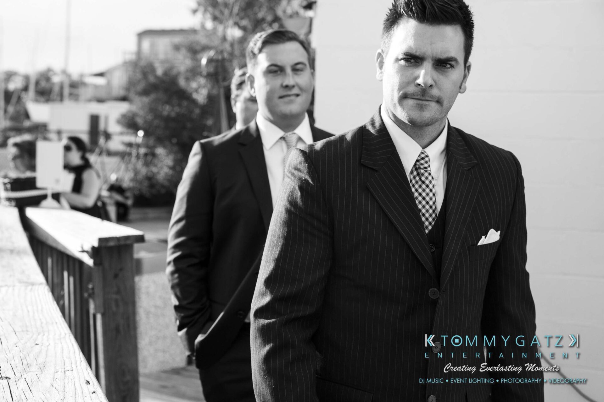 Two men in suits walk, with one in the foreground looking ahead and the other slightly behind. They are outside on a wooden walkway.