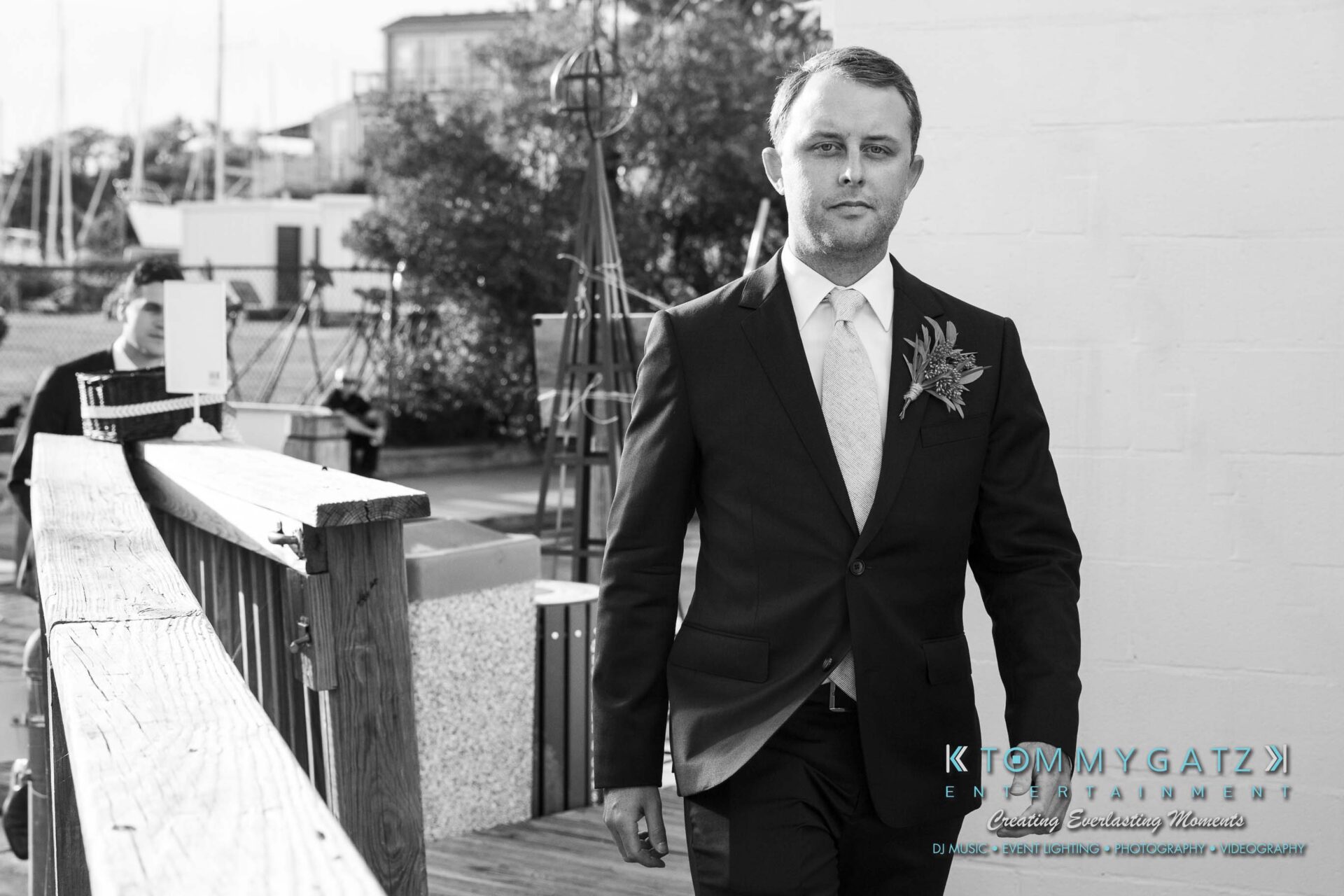 A man in a suit with a boutonniere walks on a wooden path outdoors.