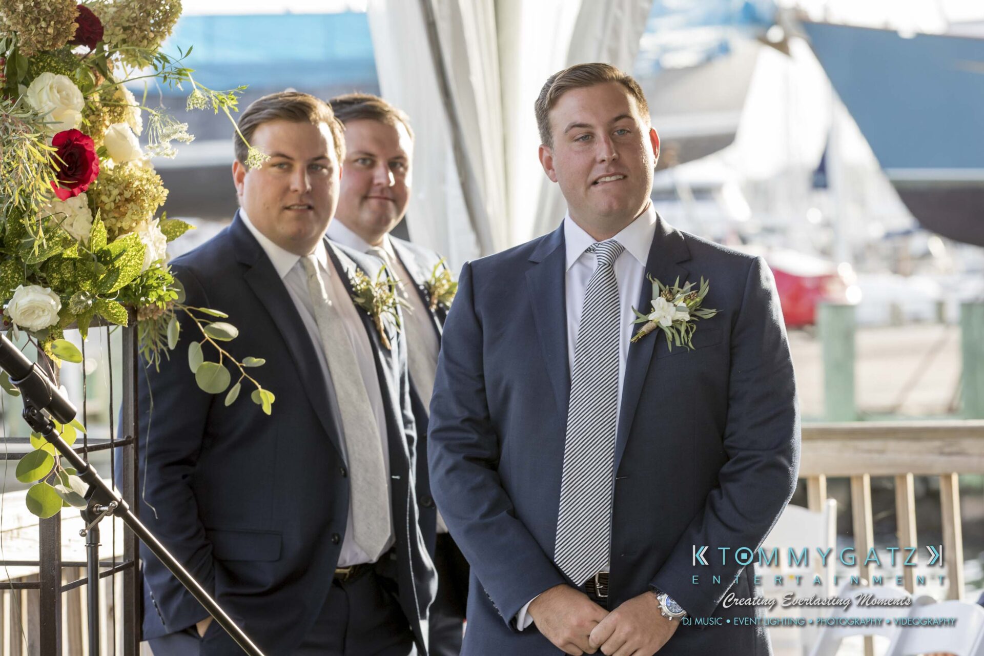 Three men in suits stand next to a floral arrangement at an outdoor event. One man is at the forefront, while two others stand nearby. Sailboats are visible in the background.