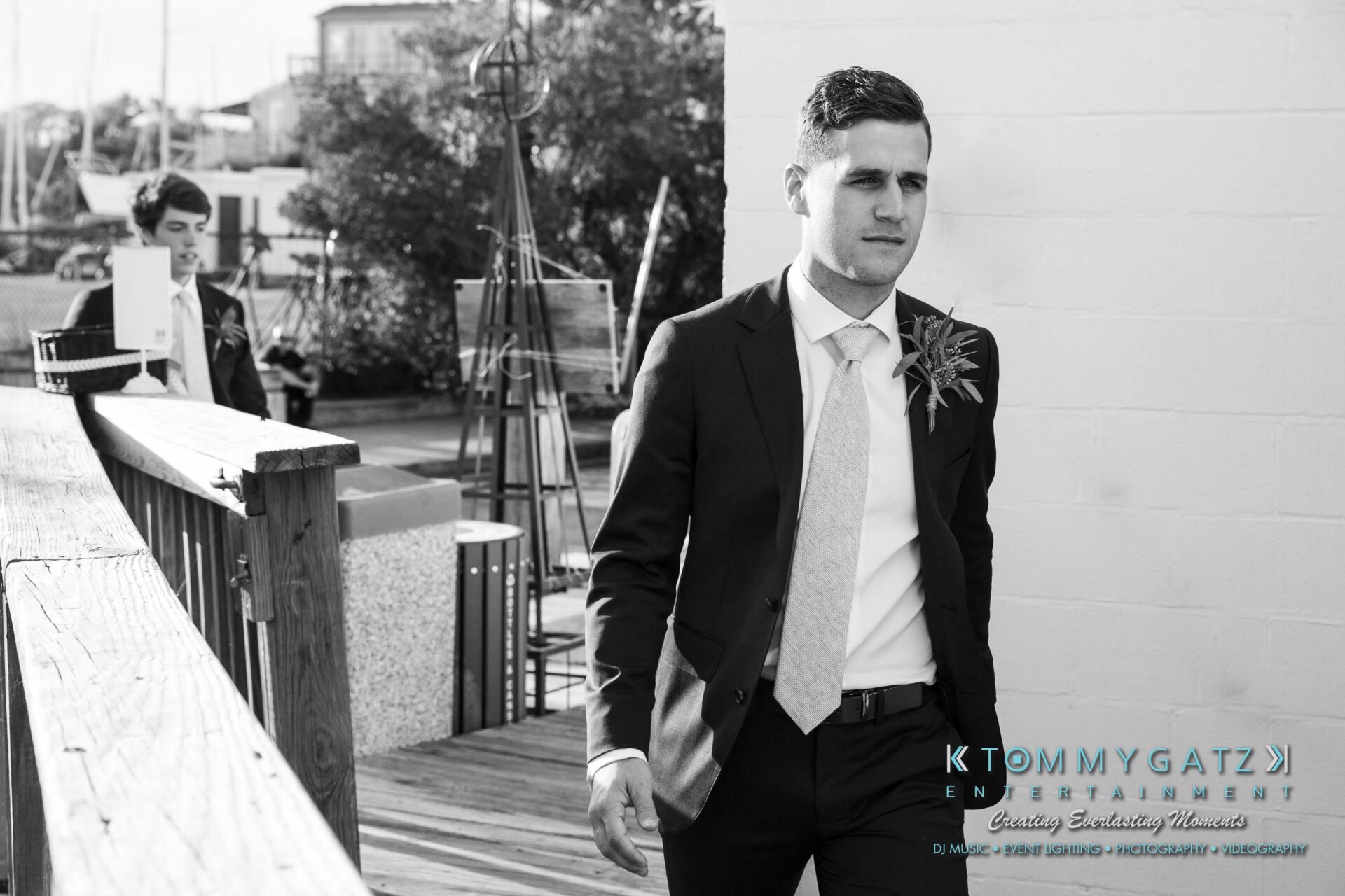 A man in a suit walks along a wooden deck, followed by another person. Buildings and a boat mast are visible in the background.