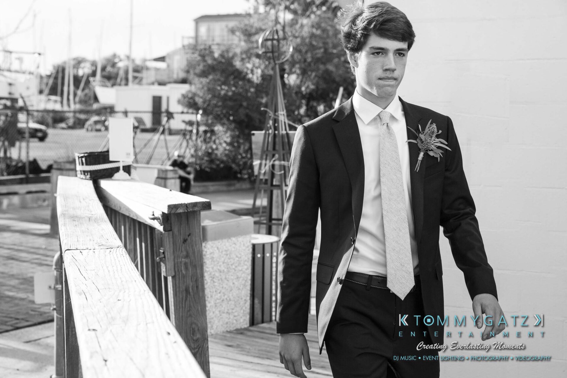 A man in a suit with a boutonniere walks on a wooden platform near boats, looking ahead.