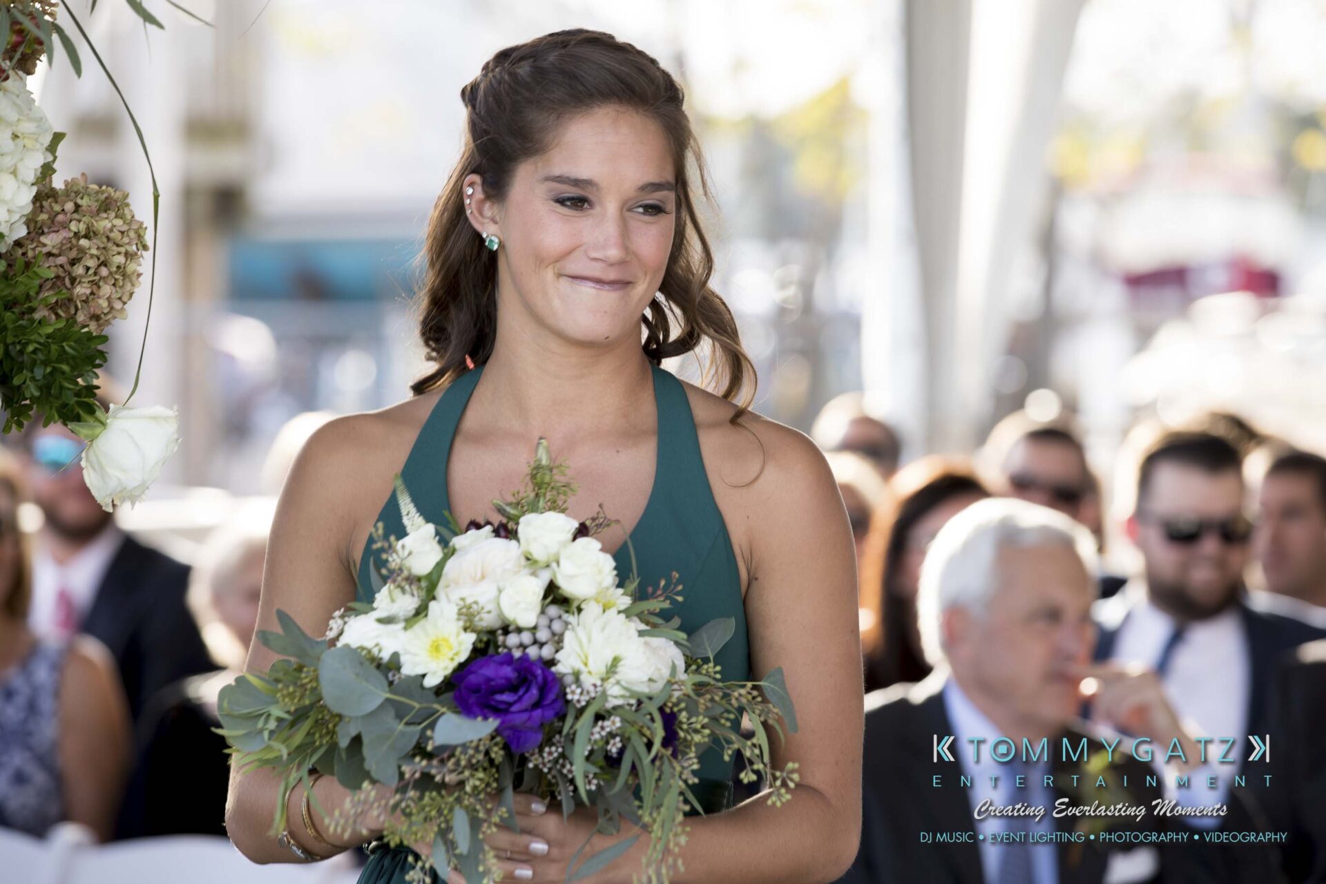 A woman in a green dress holds a bouquet of white and purple flowers while walking at an outdoor event.