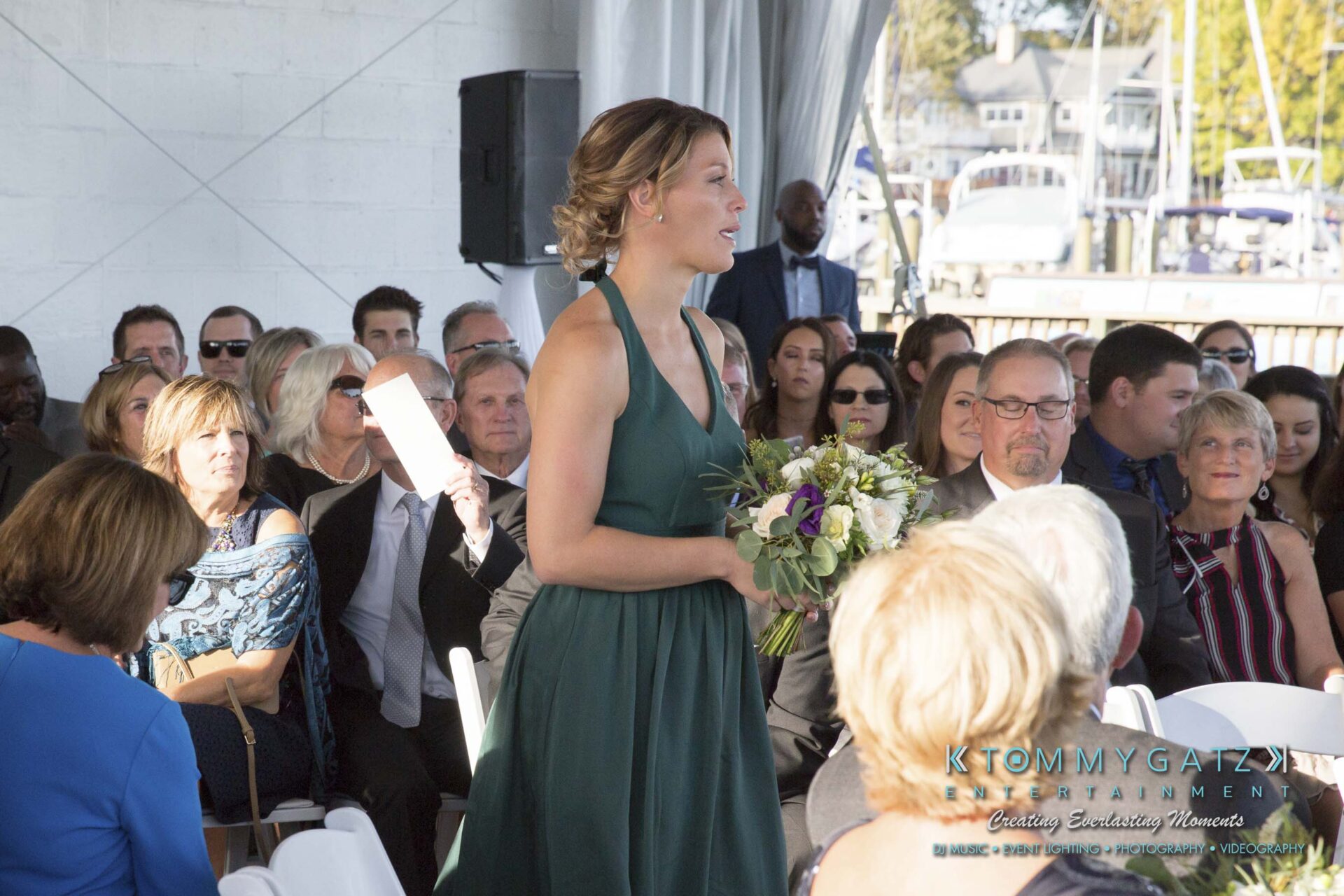 A woman in a green dress stands holding a bouquet, speaking to a seated audience at an event.