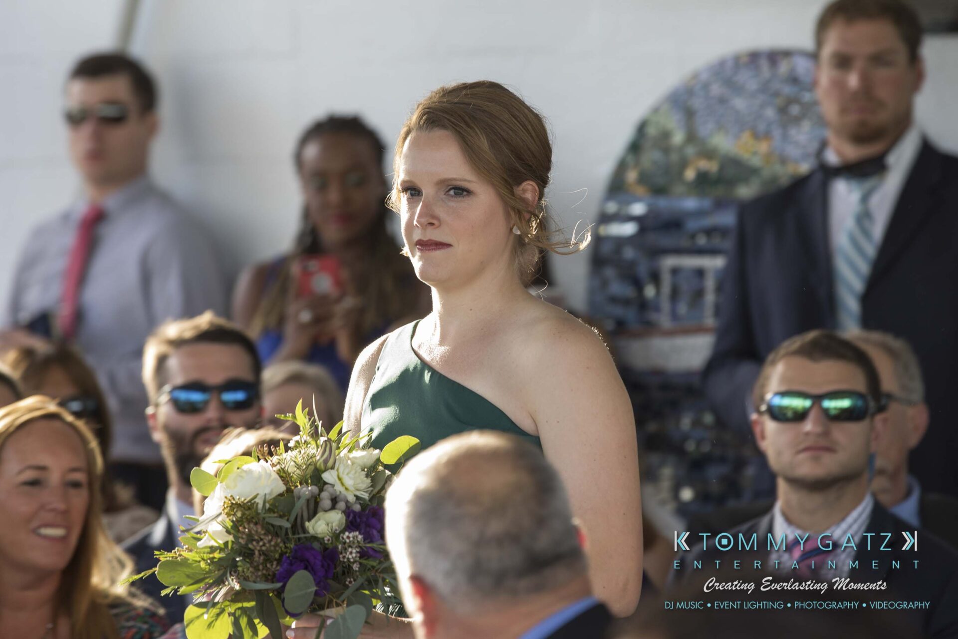 A woman in a green dress holds a bouquet while standing in front of seated people at an event.