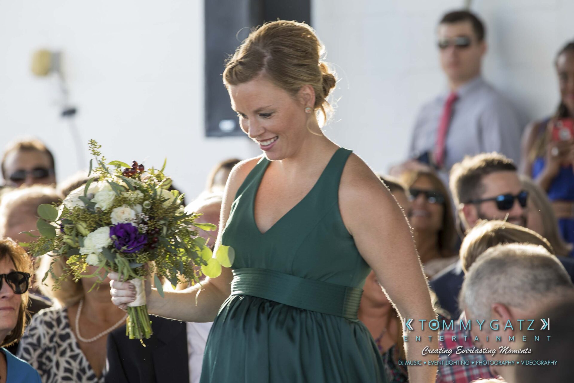 A woman in a green dress carries a bouquet of flowers while walking past seated guests at an event.