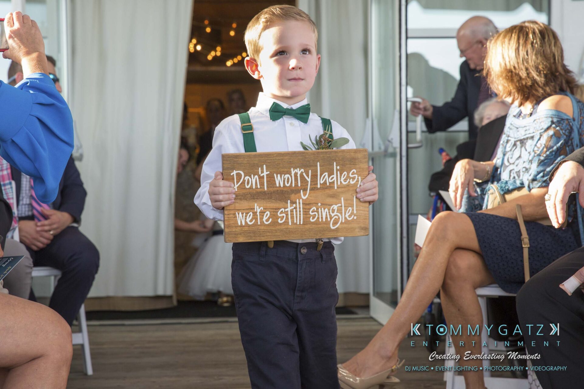 Amanda-Tyler-102117-CEREMONY-Low-Res-WM-24 - TOMMY GATZ ENTERTAINMENT A young boy in formal attire holds a sign reading, "Don't worry ladies, we're still single!" while walking down an aisle at an event.