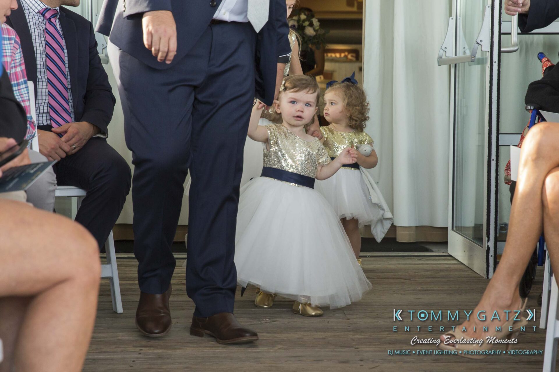 Amanda-Tyler-102117-CEREMONY-Low-Res-WM-26 - TOMMY GATZ ENTERTAINMENT Two young girls in gold and white dresses hold hands, walking beside a man in a suit at an indoor event.