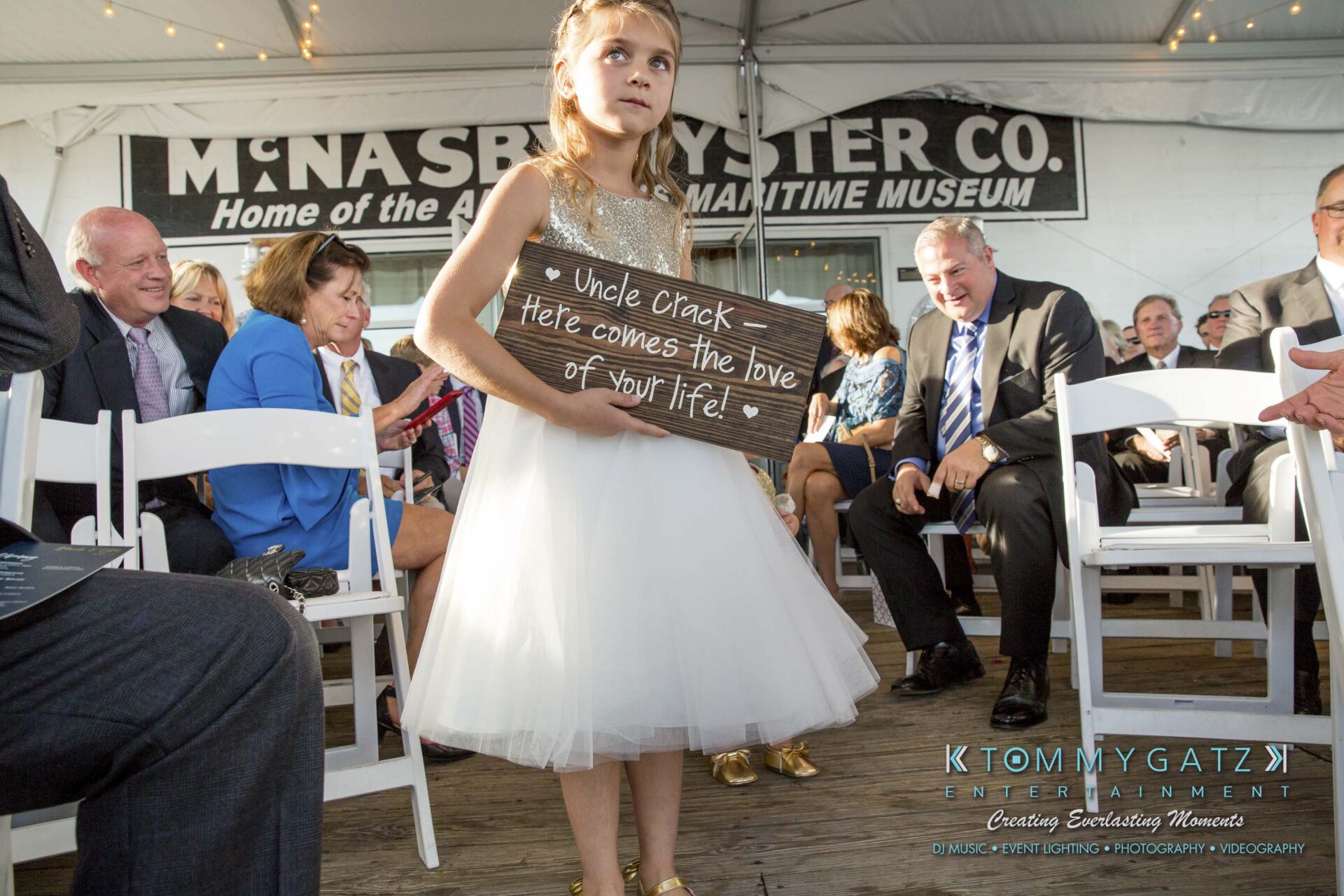 Amanda-Tyler-102117-CEREMONY-Low-Res-WM-29 - TOMMY GATZ ENTERTAINMENT A young girl in a white dress holds a wooden sign reading "Uncle Crack, here comes the love of your life" during a ceremony. People sit in rows under a tent.